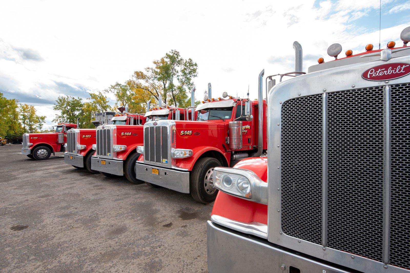 Line of red fire trucks parked on a gravel lot with trees and a partly cloudy sky in the background.