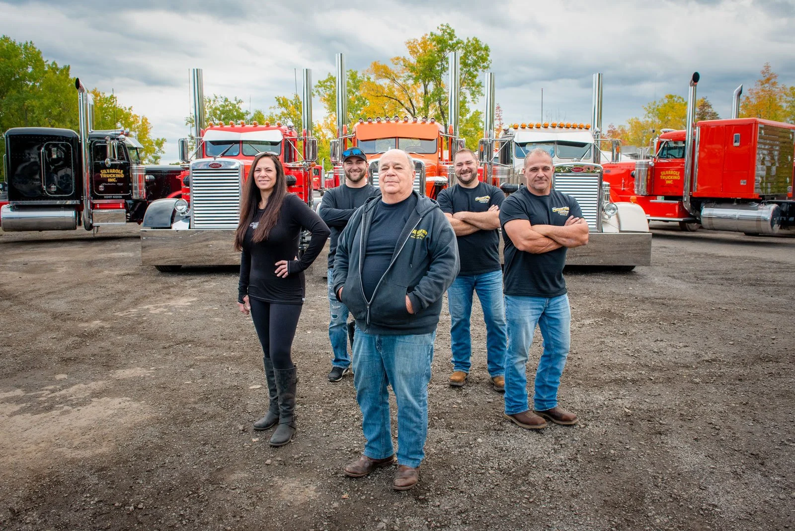 Group of five people standing in front of large red fire trucks outdoors during daytime with cloudy sky and trees showing fall foliage.