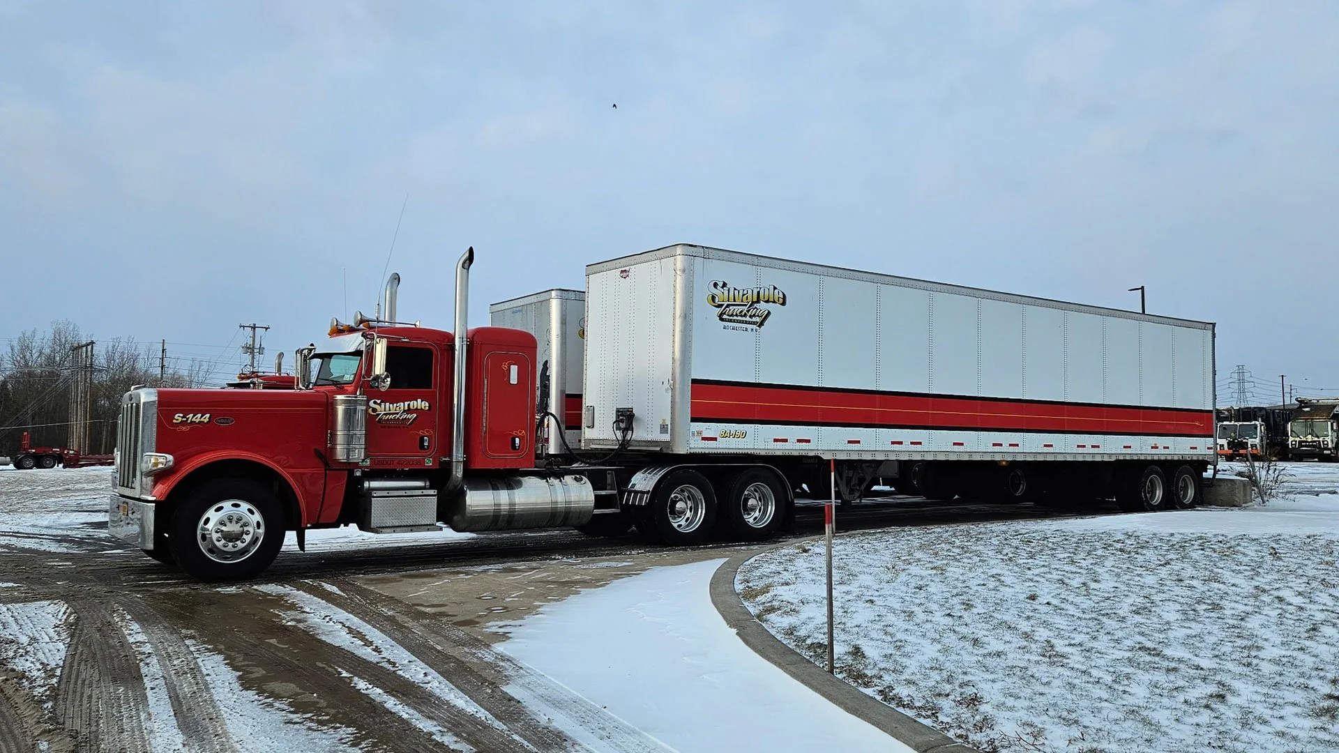 A red semi-truck with a white trailer parked on a snowy ground, with a cloudy sky overhead and a few distant vehicles in the background.