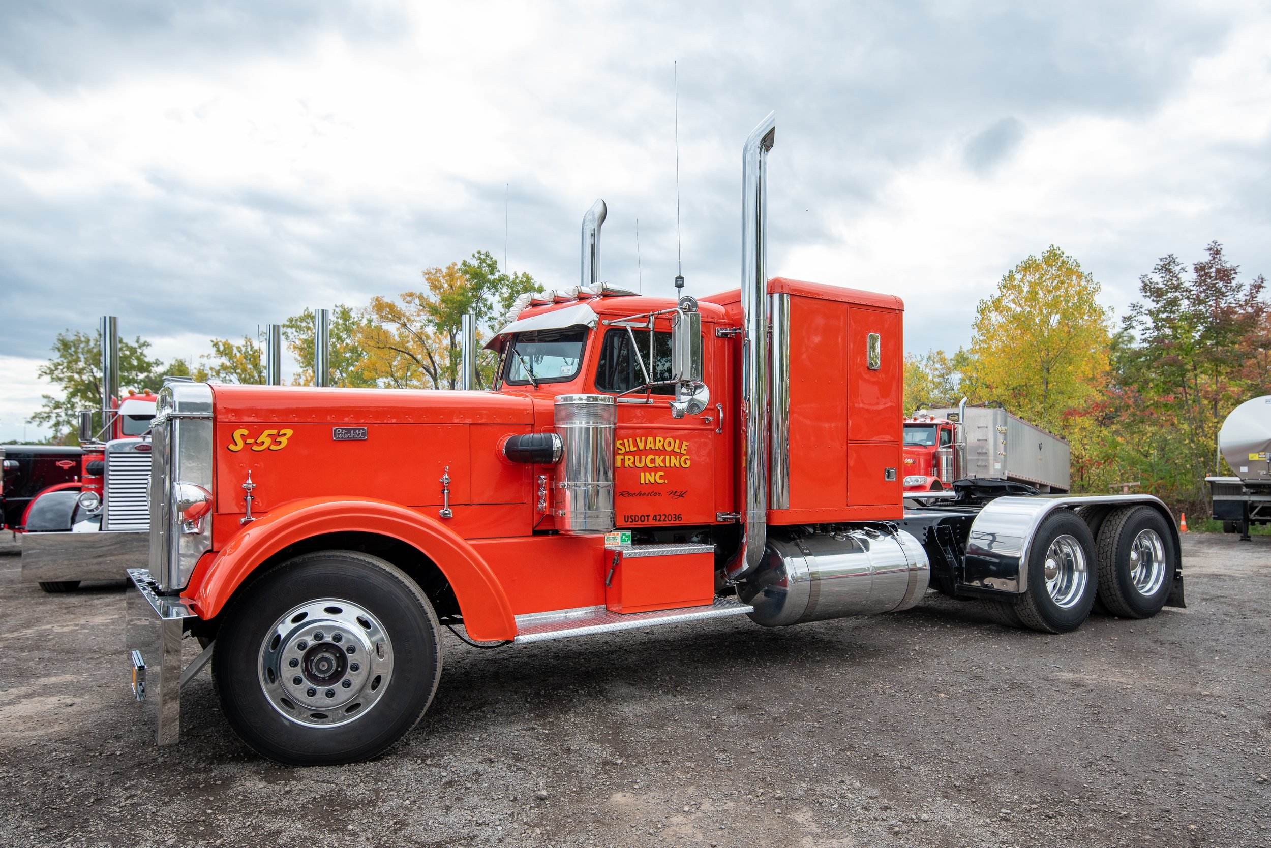 Red semi-truck with chrome exhaust stacks and grooming orange paint parked on a dirt lot with trees in the background.
