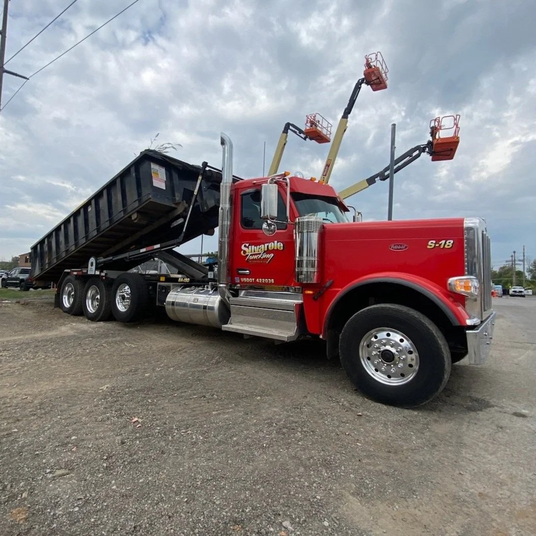 Red dump truck with a raised black bed and three lift arms with orange buckets, parked on a dirt lot under cloudy skies.