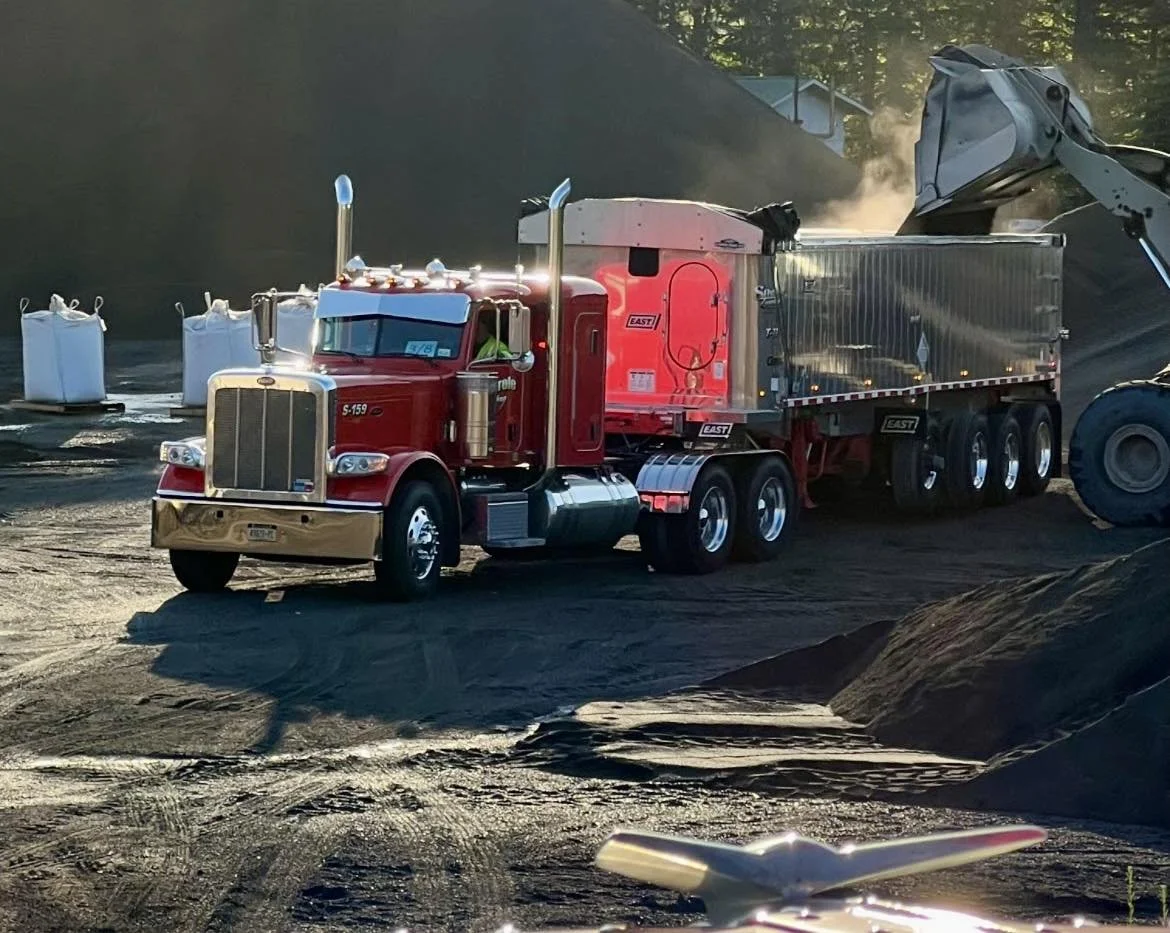 A large red semi-truck dumping a load of black dirt or coal at a construction site during daylight. There are white bags filled with materials in the background.