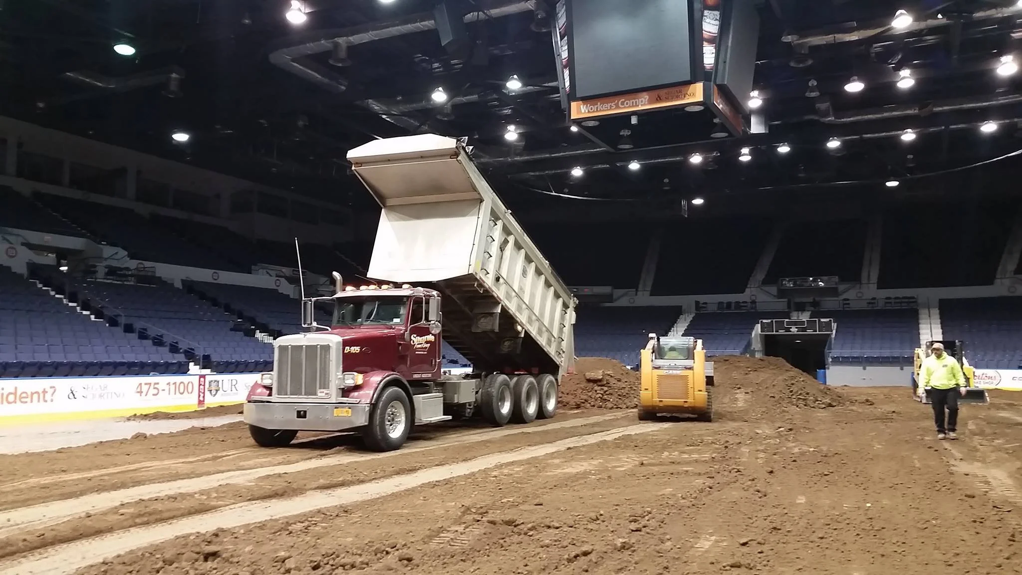 Indoor arena under construction with a Dump truck, soil compactor, and construction workers on the dirt floor.