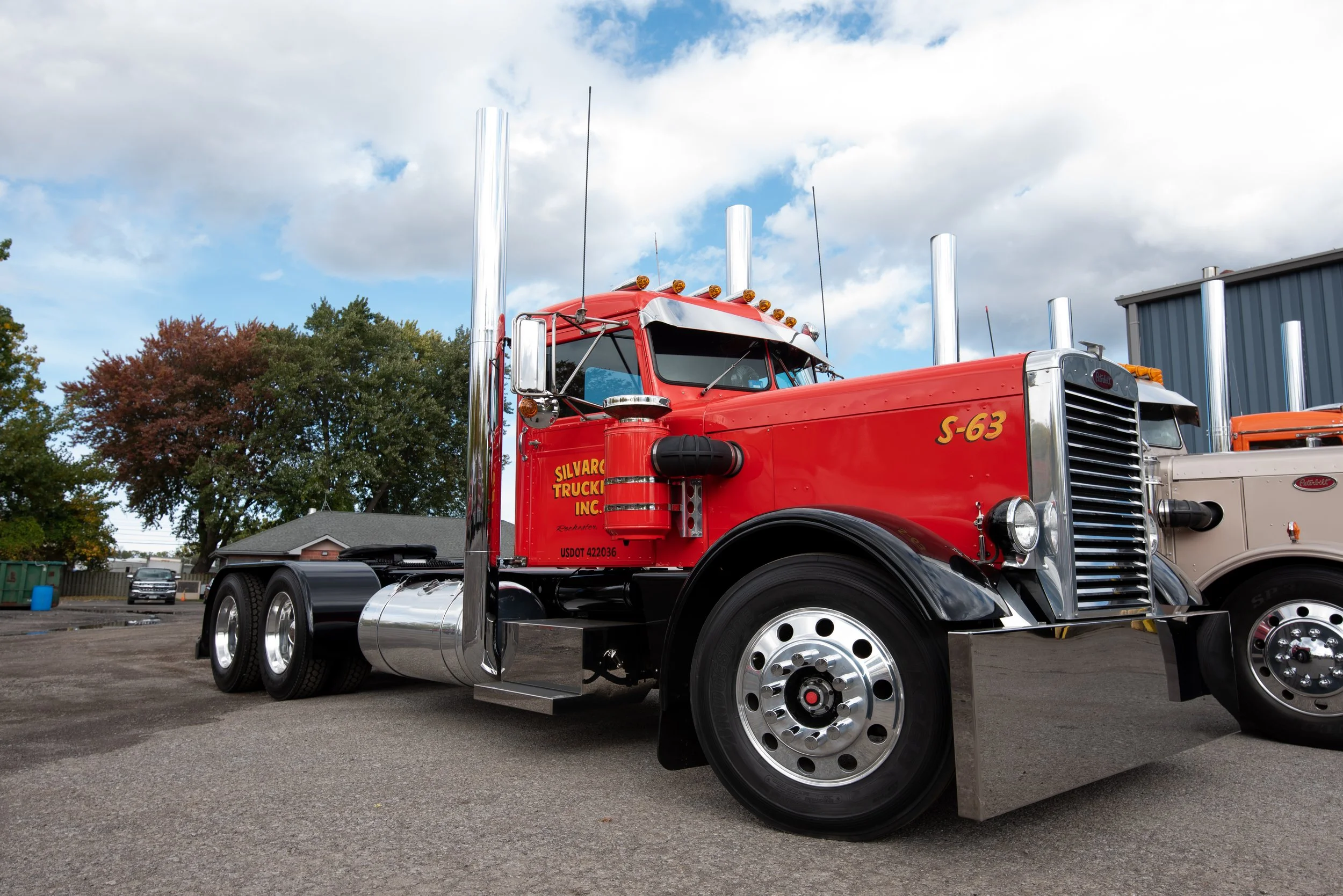 Red semi-truck parked in an outdoor lot with trees and a building in the background, under a partly cloudy sky.