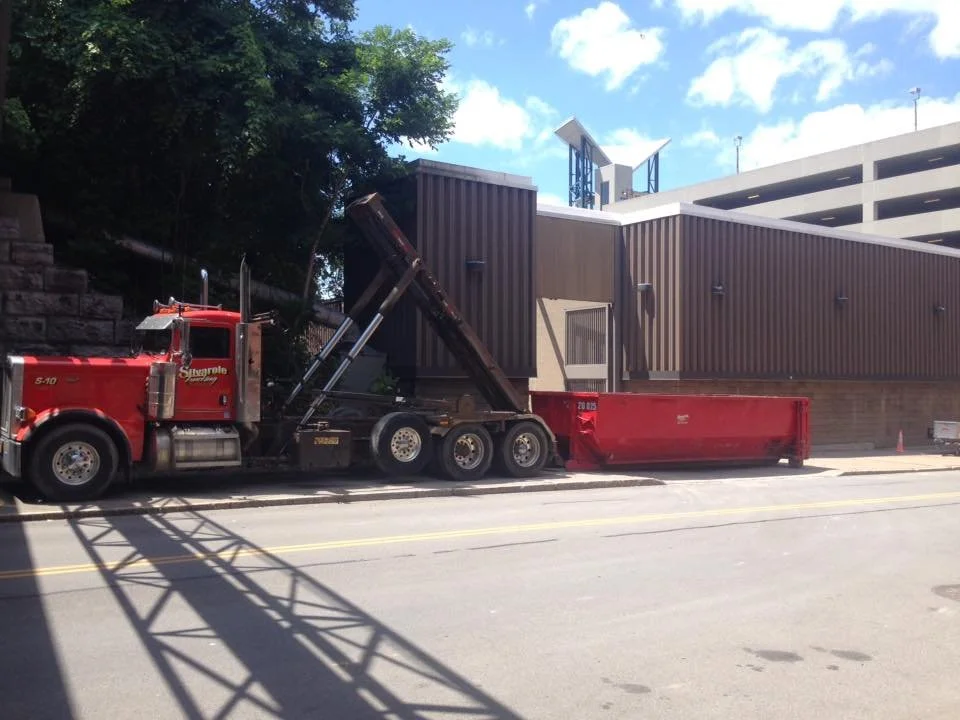 A red dump truck with its bed lifted, parked on the street next to a building with brown metal siding and a parking garage in the background.