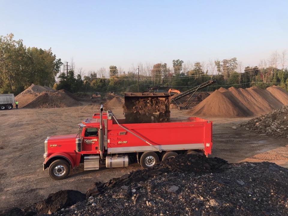 Construction site with a red dump truck in the foreground and gravel piles, an excavator, and a conveyor belt in the background.