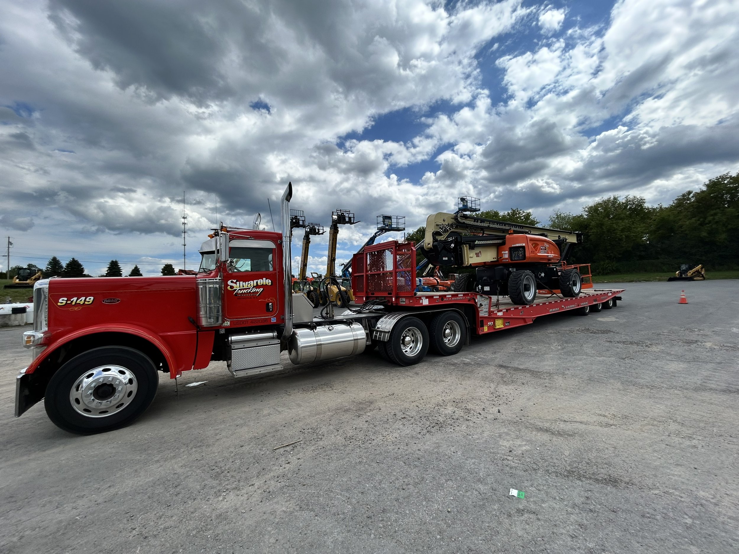 A red truck carrying construction or industrial equipment, including a large machine with an extendable arm, parked on a gravel lot under a partly cloudy sky.