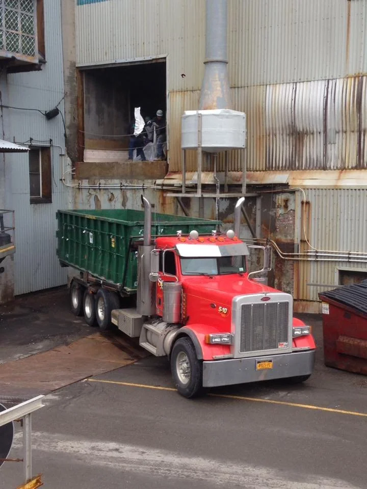 A red semi-truck parked in an industrial area with a large green container in the trailer. Two workers are inside a warehouse, visible through an opening in the corrugated metal building.