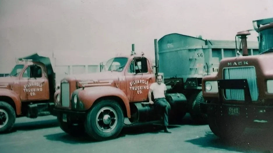 A man standing next to vintage trucks with the label "Silvarole Trucking Co." on the side, parked on a lot.