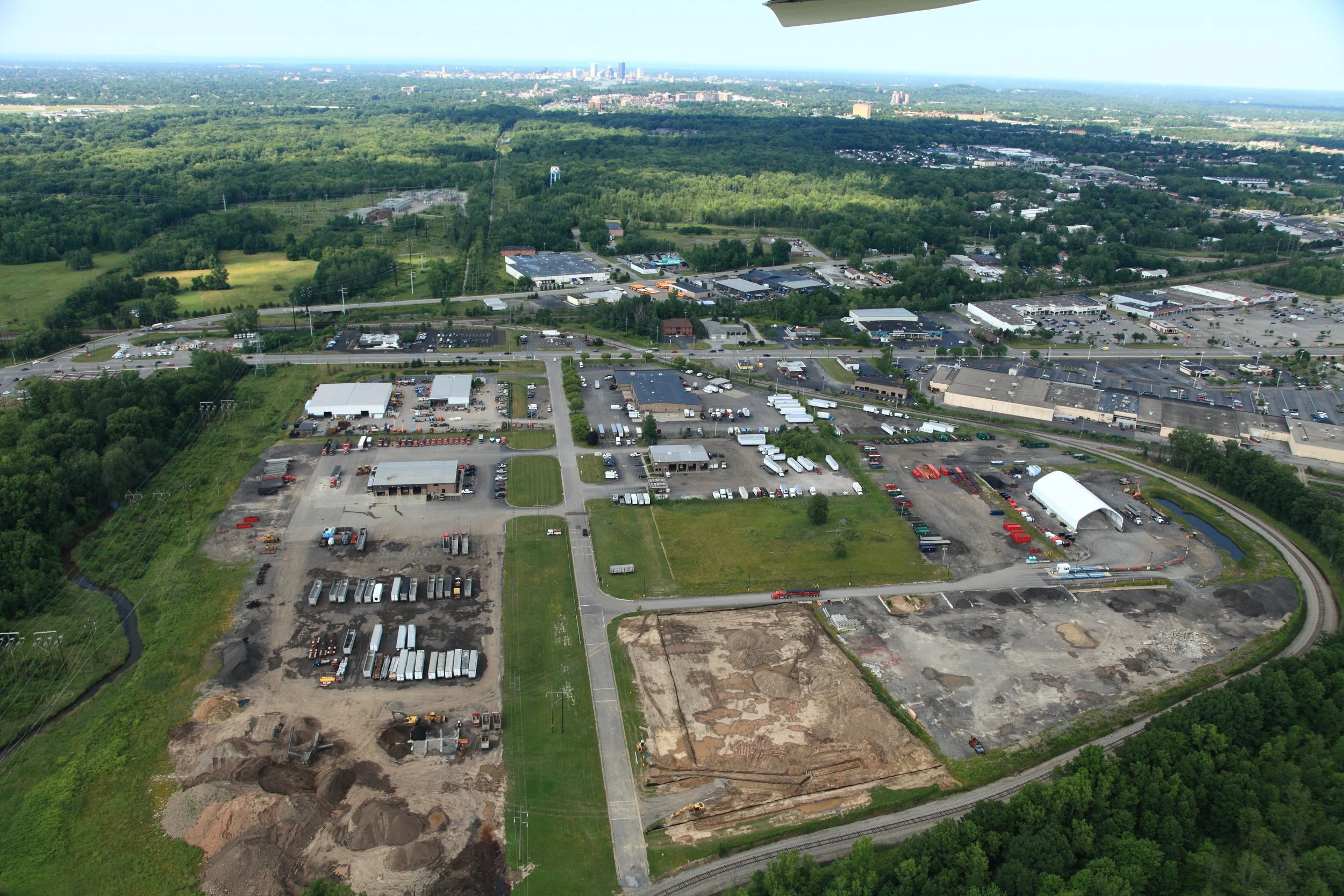 Aerial view of a mix of commercial and industrial areas with parking lots, warehouses, construction sites, surrounded by lush green trees and a distant city skyline under a clear sky.