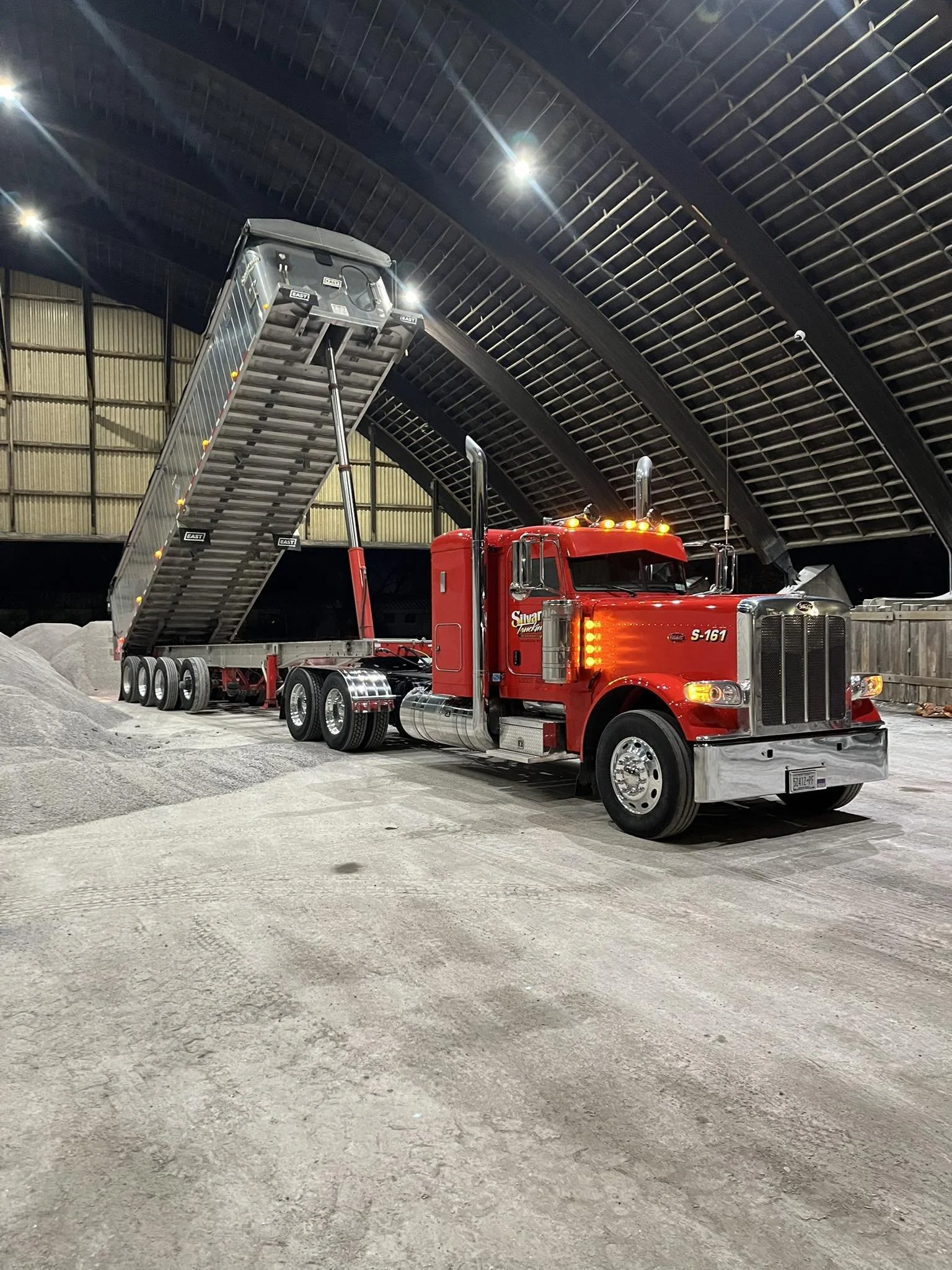 A red dump truck inside a large, indoor storage facility, with its raised bed holding a load of gravel or rocks.