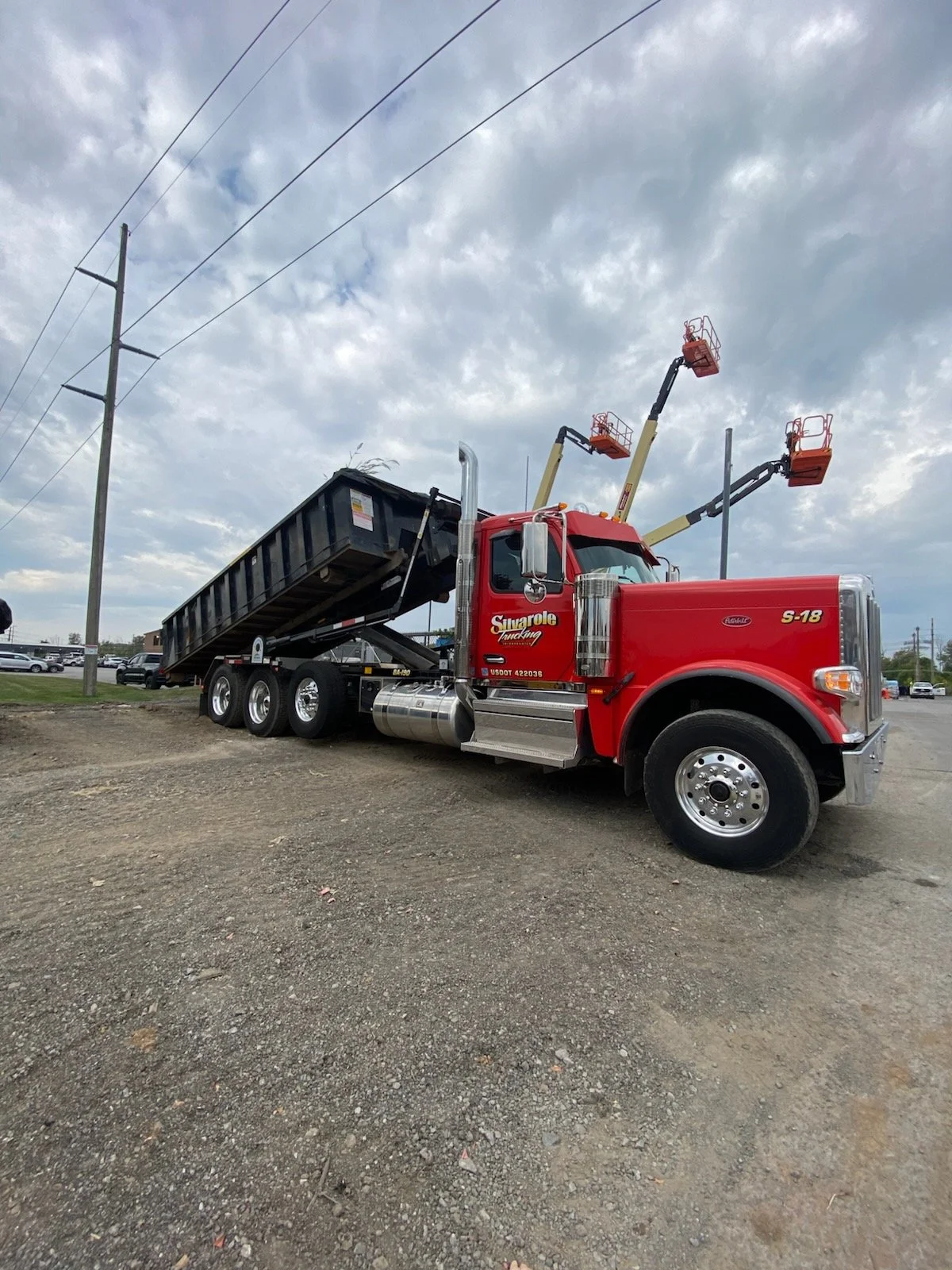 A red dump truck with a black bed is parked on a gravel lot. The truck has the words "Sunshine Trucking" written on the cabin. The truck is equipped with multiple cherry pickers extending upwards, with baskets at the ends for lifting workers. Overhead power lines are visible above, and the sky is cloudy.