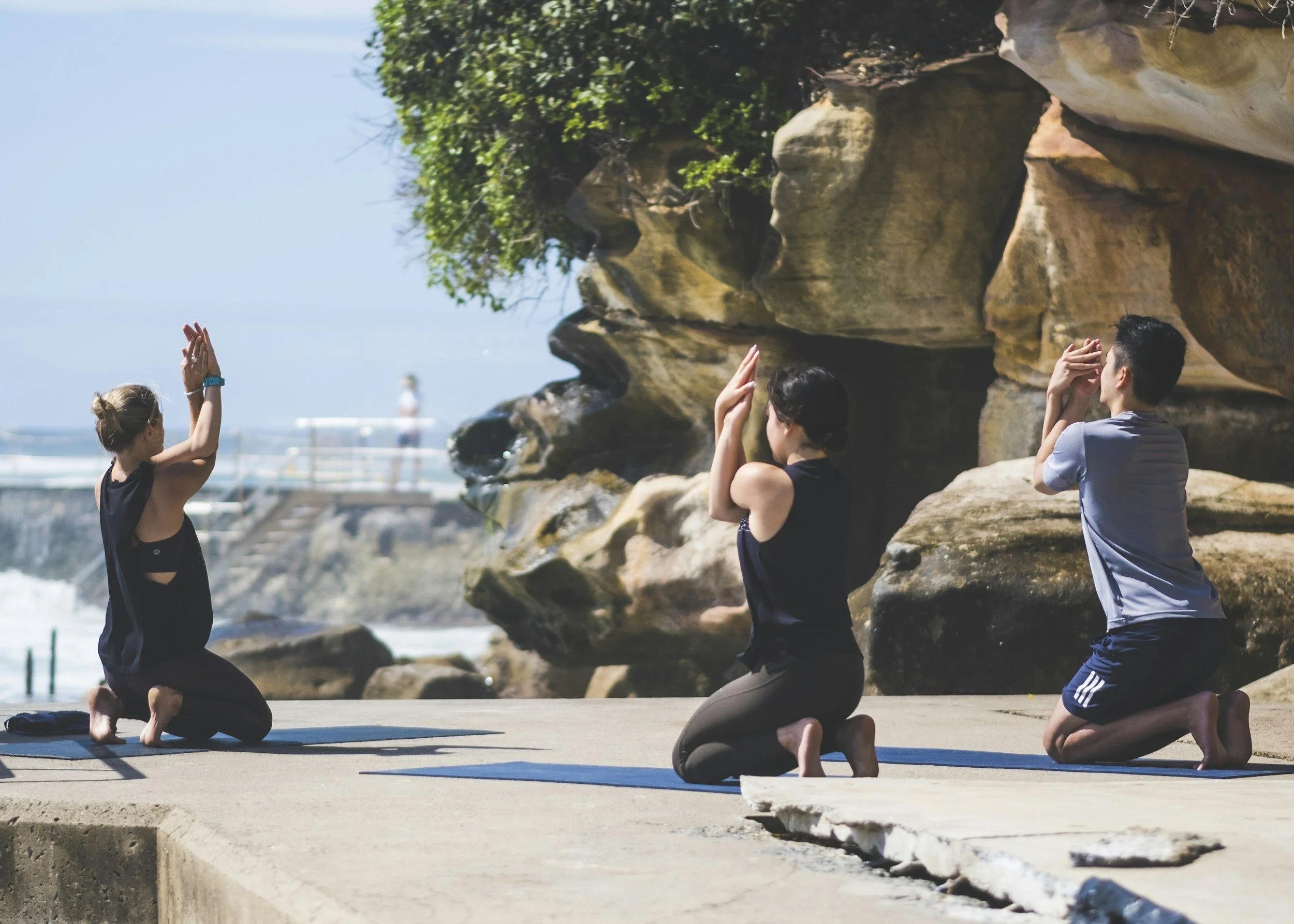 Three people practicing yoga outdoors on a sunny day near rocks, possibly by the beach, with a woman in a black top and two others, all in kneeling positions with hands raised in prayer gesture.