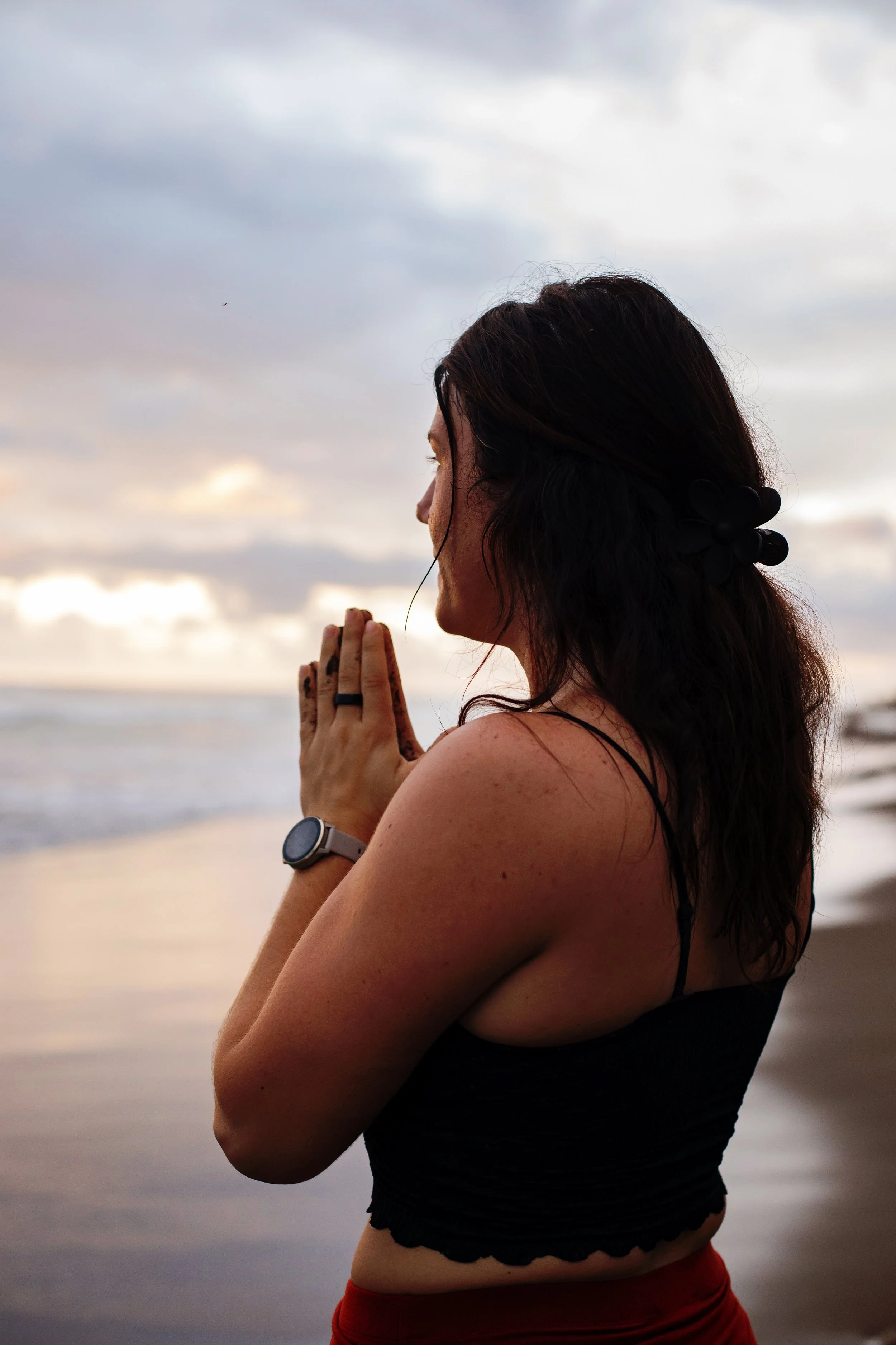 A woman practicing yoga on a beach during sunset, standing with her hands in prayer position and eyes closed.