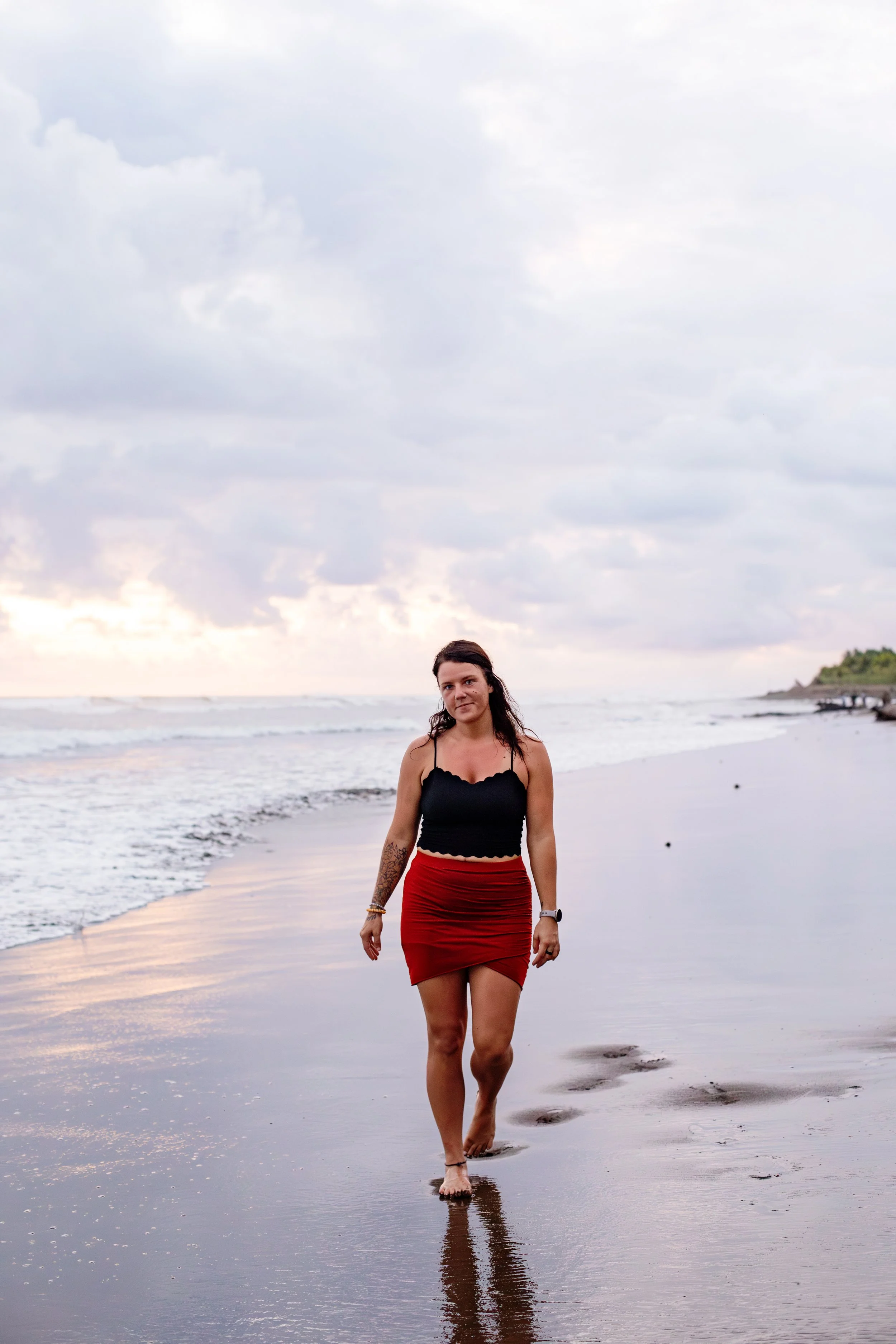 A young woman with dark hair and tattoos on her arm walking barefoot along the shoreline at sunset or sunrise, wearing a black top and a red skirt, with a cloudy sky and a distant landmass in the background.