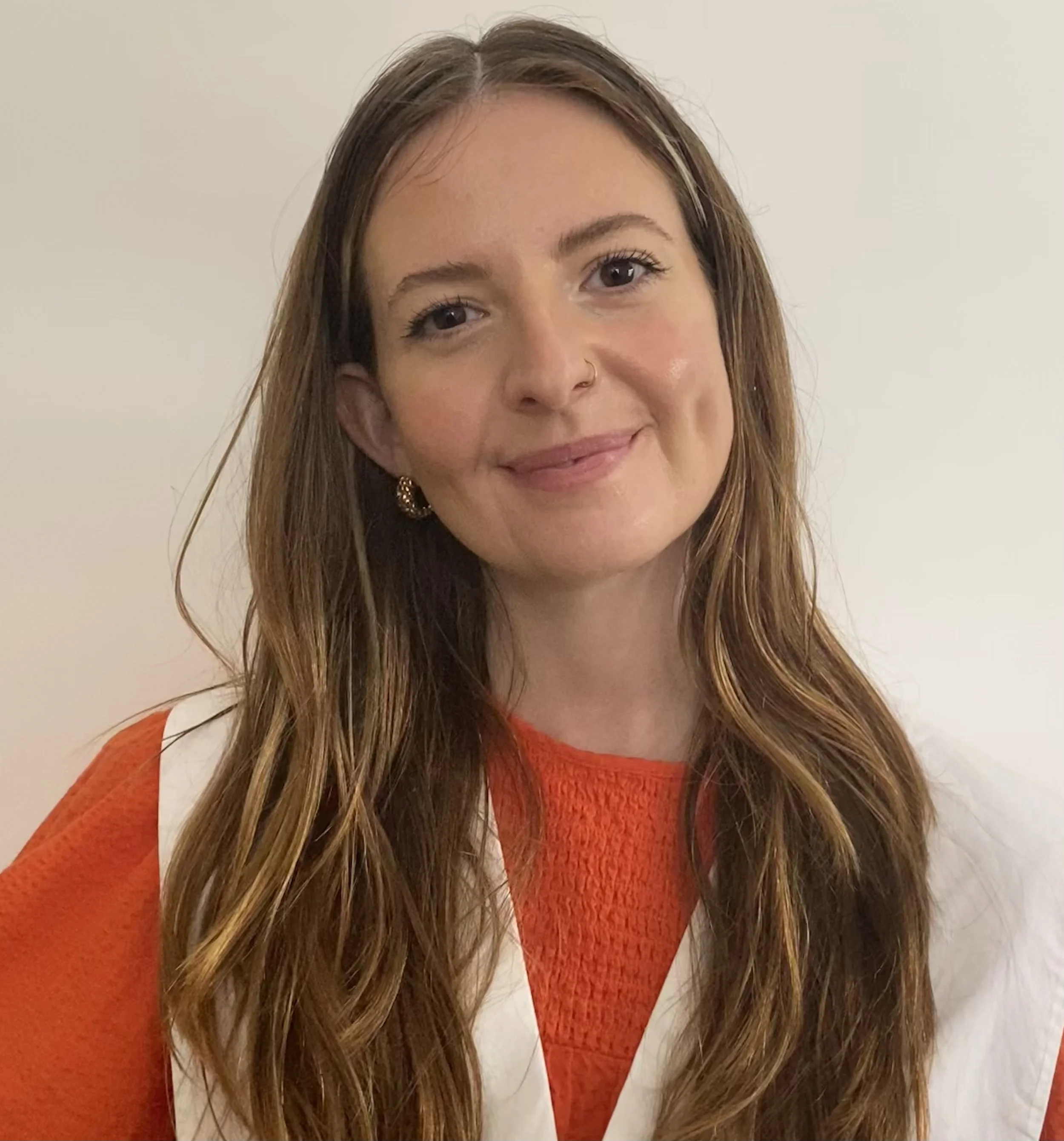 A young woman with long, wavy brown hair, wearing a red sweater and gold earrings, smiling against a plain white background.