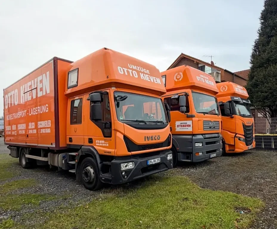 Drei orangefarbene Umzugstransporter von Otto Kievet stehen auf einer Wiese vor Häusern.