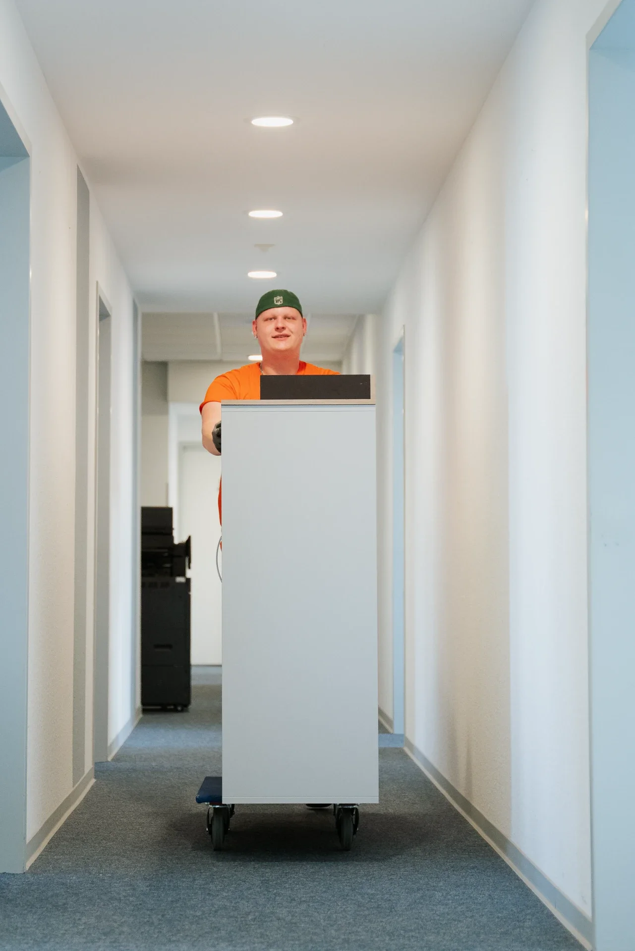 Ein Mann in orangefarbenem T-Shirt schiebt eine leere, weiße Kiste auf Rollwägen durch einen Flur in einem Bürogebäude.