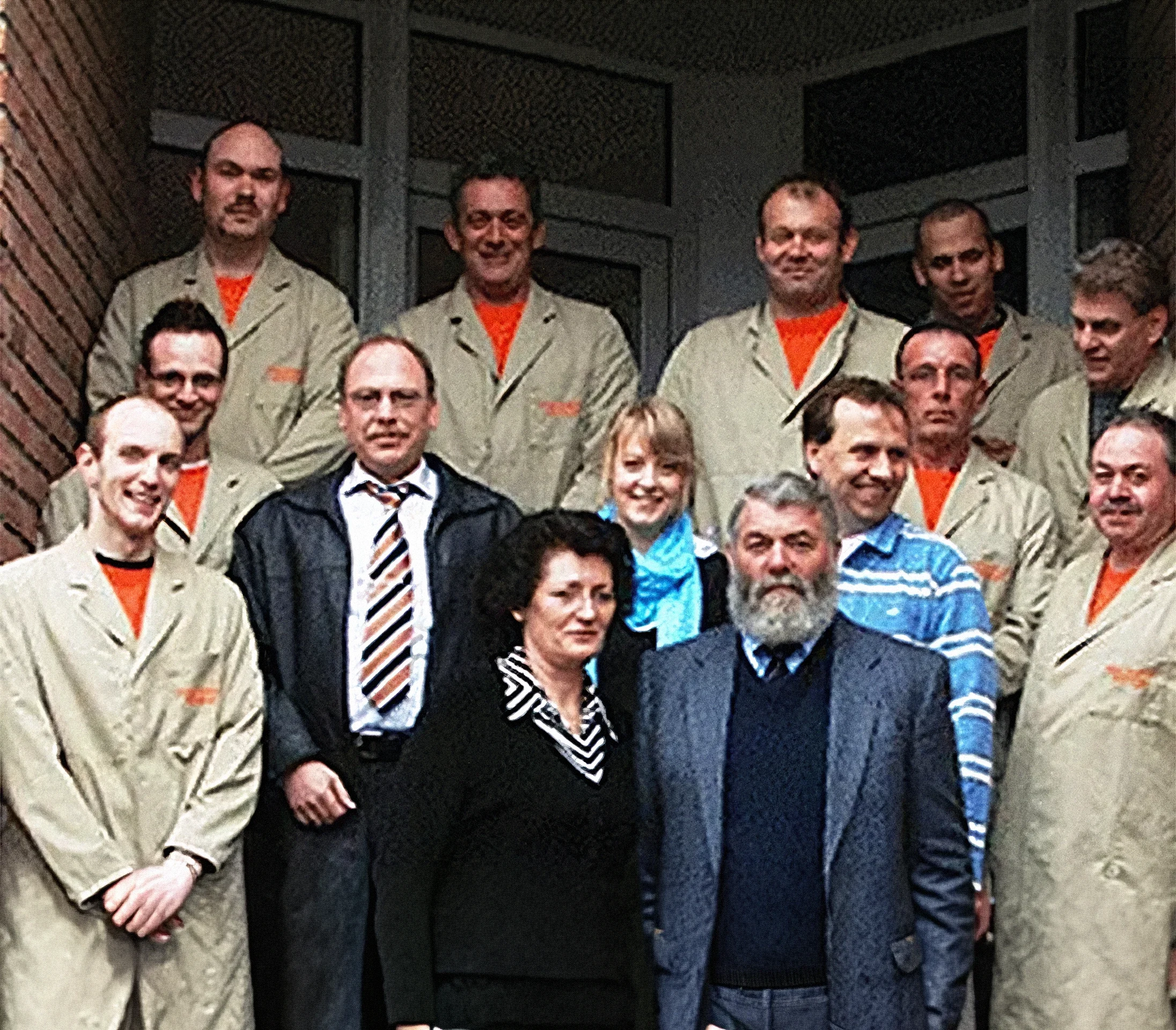 Gruppe von Männern und Frauen, einige in beige Arbeitsjacken, einige in Anzügen, vor einem Gemäuer. Es handelt sich vermutlich um eine Arbeits- oder Teamsitzung.