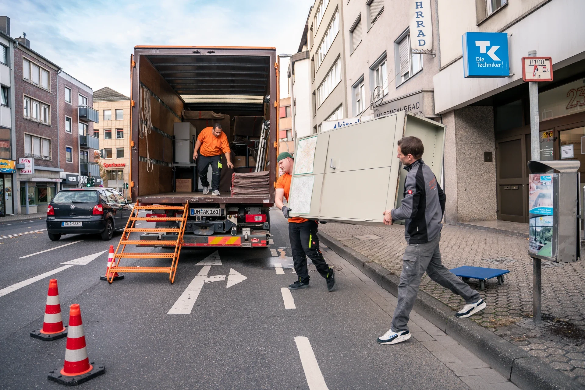 Drei Männer laden Möbel in einen Lieferwagen, der auf einer Straßenmitte parkend steht. Sie tragen eine Couch und eine Schrankwand. Die Orangefarbenen Tragehandschuhe, die sie tragen, deuten auf eine professionelle Umzugsarbeit hin.