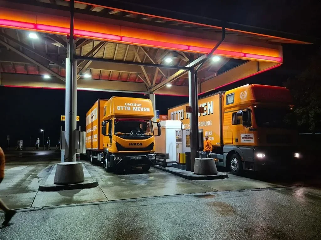 Zwei orangefarbene Umzugsfahrzeuge an einer Tankstelle bei Nacht, die für einen Umzug vorbereitet werden.