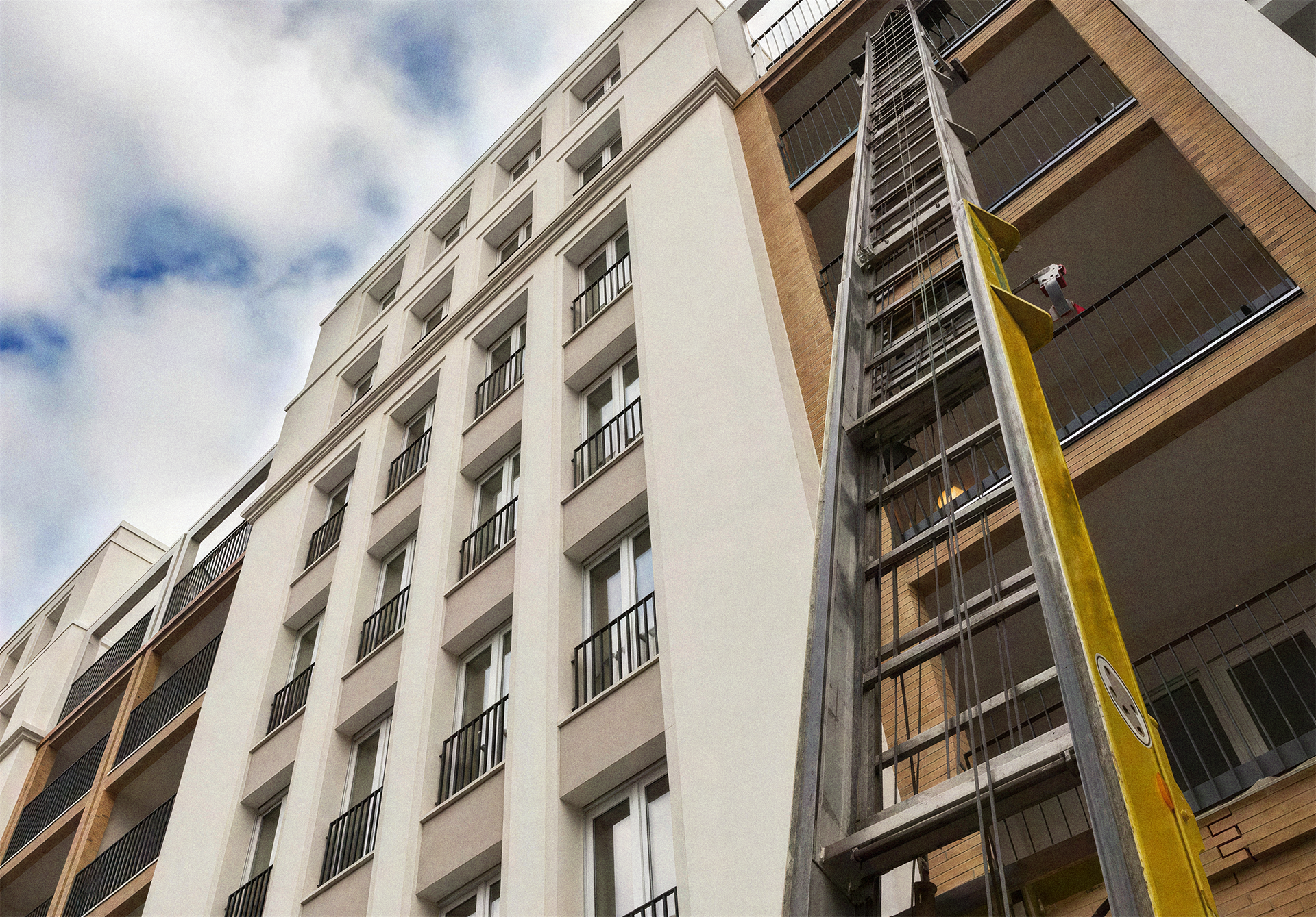 Aufzugschacht wird an einem modernen Mehrfamilienhaus installiert, sichtbar auf einem Balkon in der Nähe, mit Wolken am Himmel im Hintergrund.