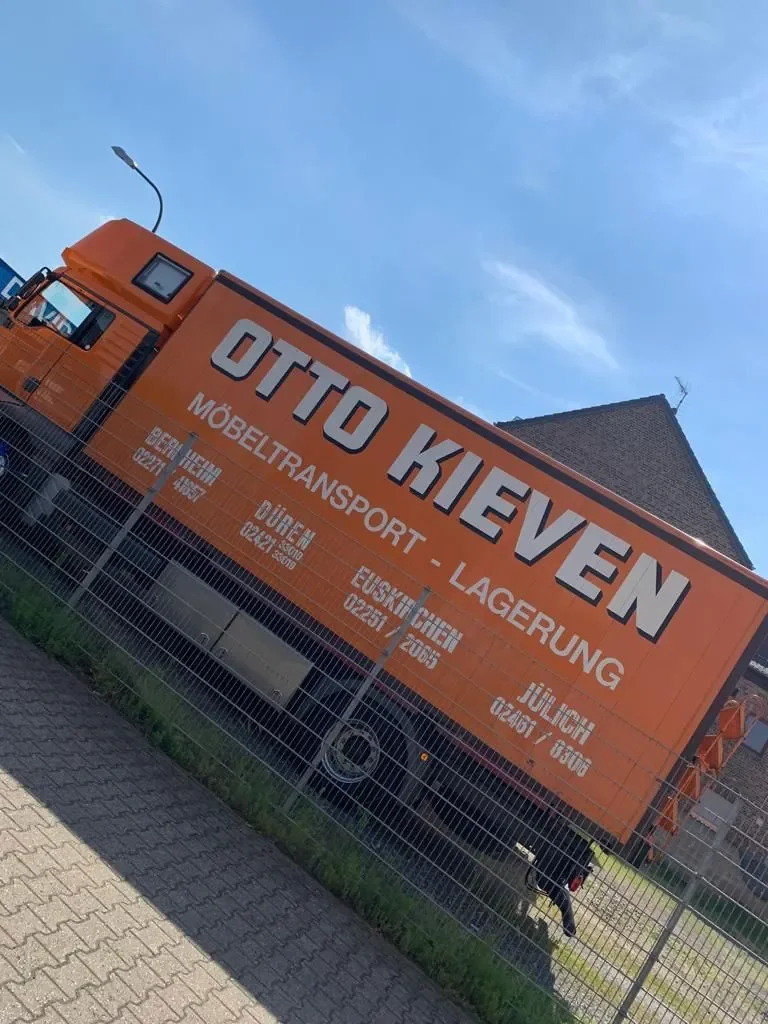 Ein orangefarbiger LKW mit dem Schriftzug 'OTTO KIEVEN Möbeltransport - Lagerung' steht hinter einem Metalzaun.