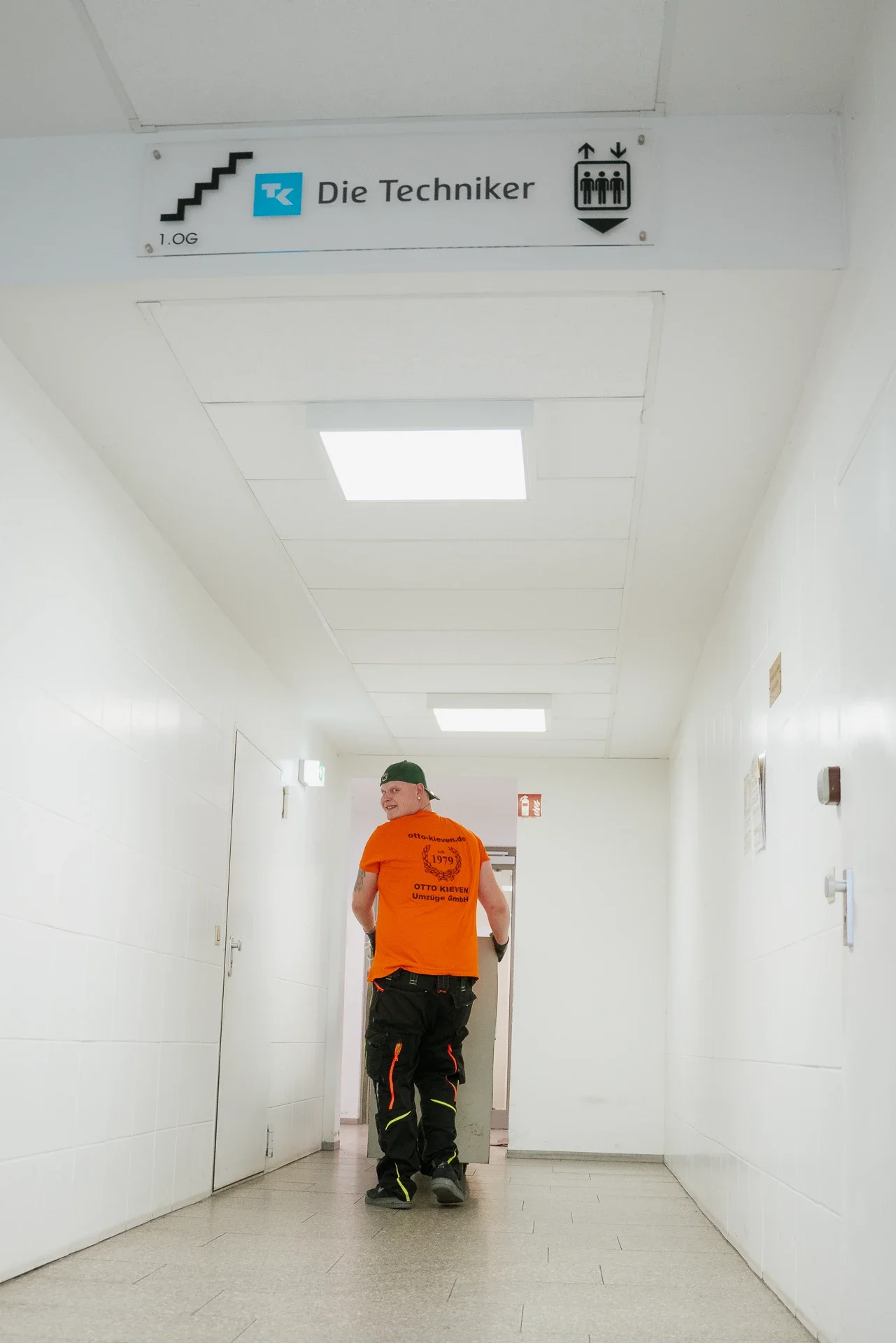 Ein Mann in schwarz-orange Arbeitskleidung und grünem Hut läuft in einem weißen Flur. Über ihm hängt ein Schild mit dem Text 'Die Techniker' und einem Aufzugsymbol.