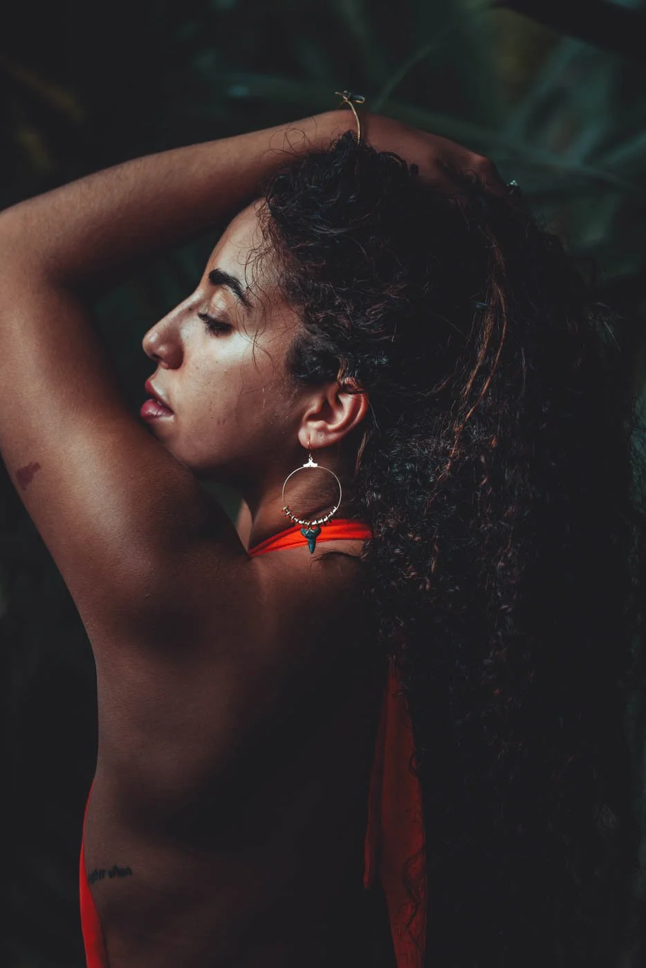 A woman with curly dark hair and earrings, posing with her arm behind her head, in a dark, leafy environment.