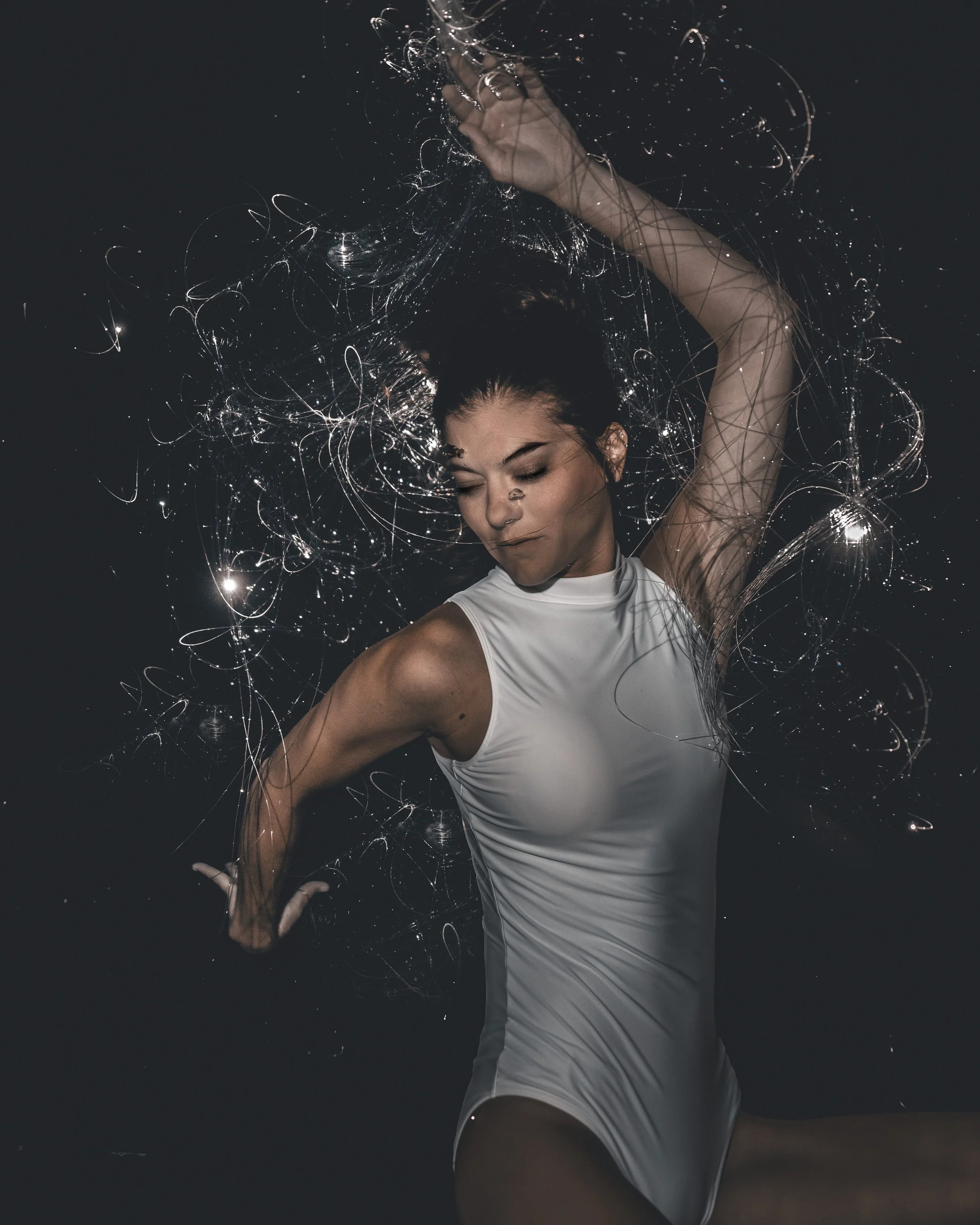 A woman in a white sleeveless leotard with dark hair tied up, appears to be dancing with liquid-like light streaks surrounding her against a dark background.