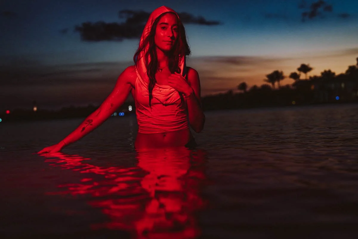 A woman wearing a sleeveless top standing waist-deep in water during sunset, with a dark sky and some trees in the background, illuminated by red and blue lighting.