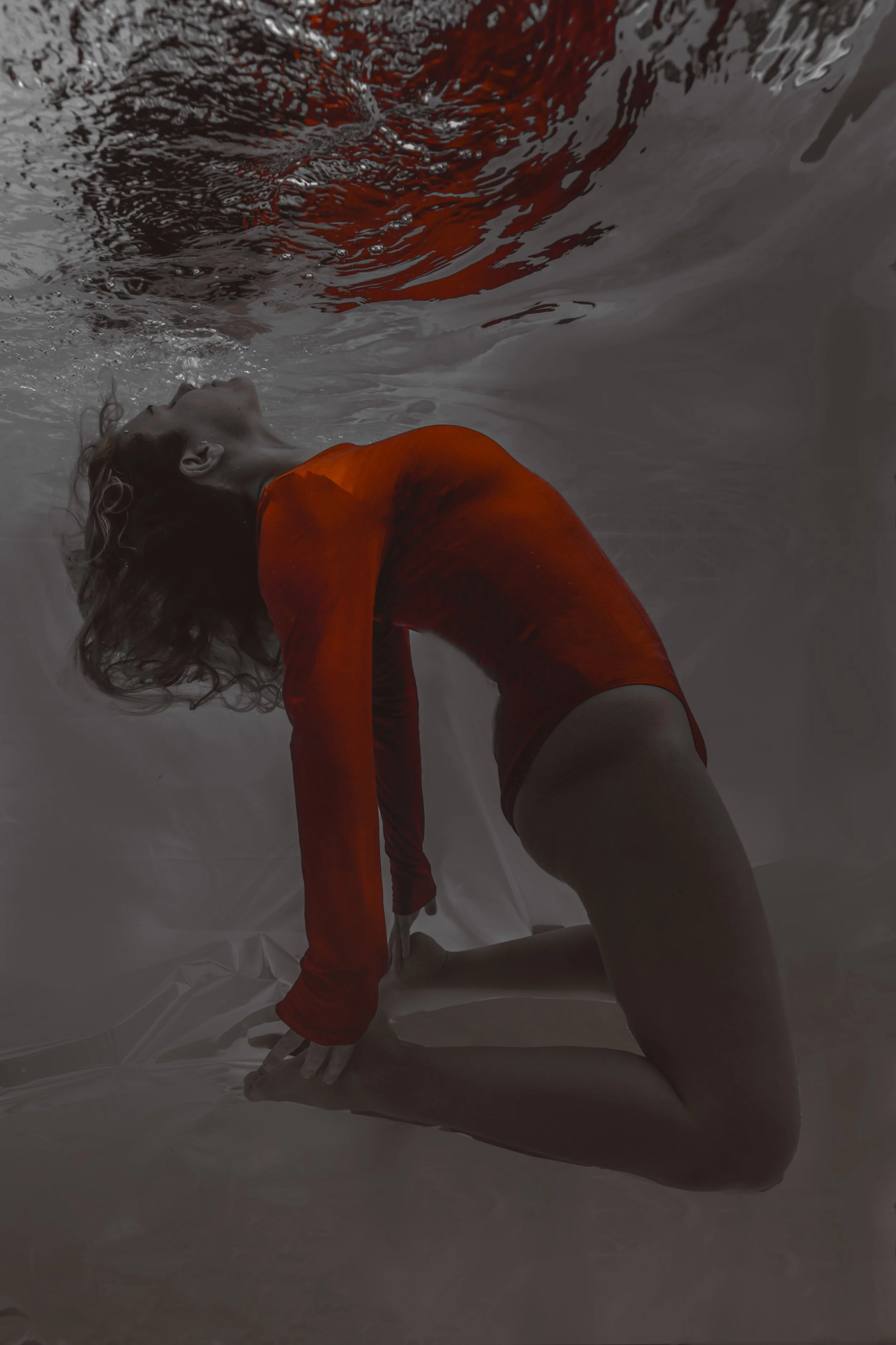 A woman in a red long-sleeve leotard is kneeling on the floor and tilting her head back while in the water, with her hands resting on her feet.