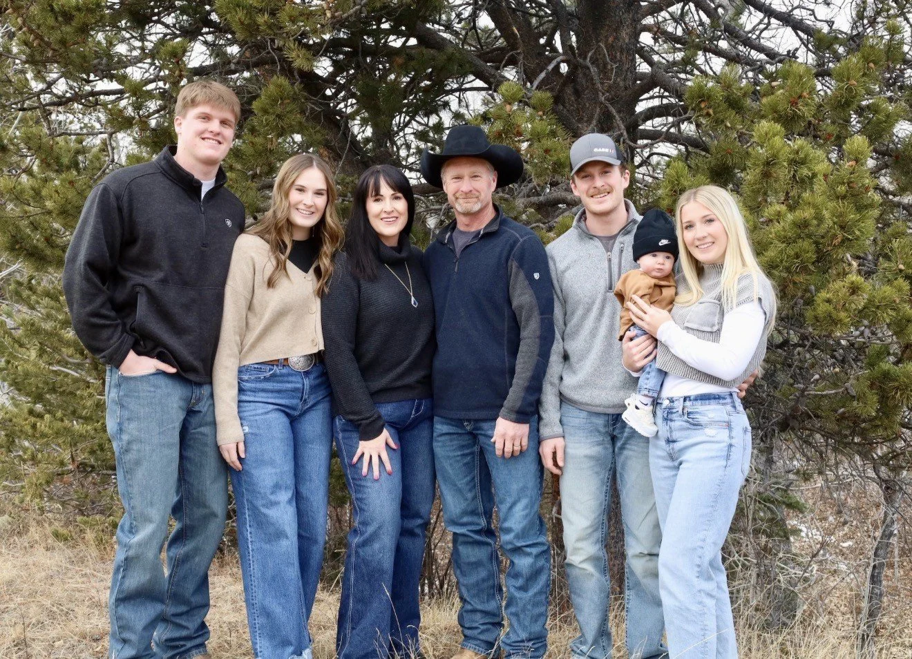 A family of seven standing outdoors in front of a tree, smiling at the camera. The family includes five adults, one of whom is holding a baby. They are dressed casually in jeans and jackets.