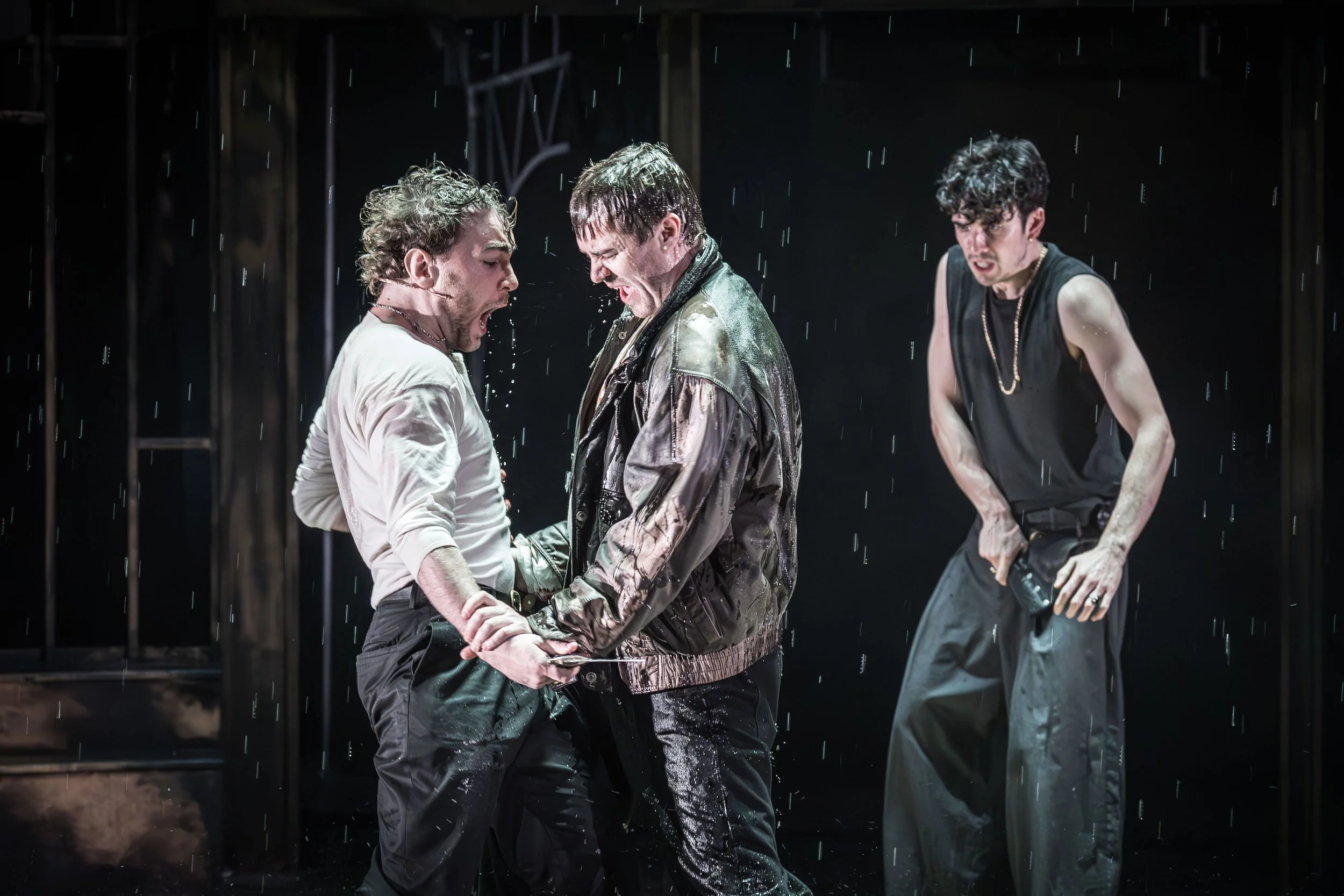 Three men in casual clothing are standing in the rain on a dark stage, with two of them looking intensely at each other while the third observes with a grimace.