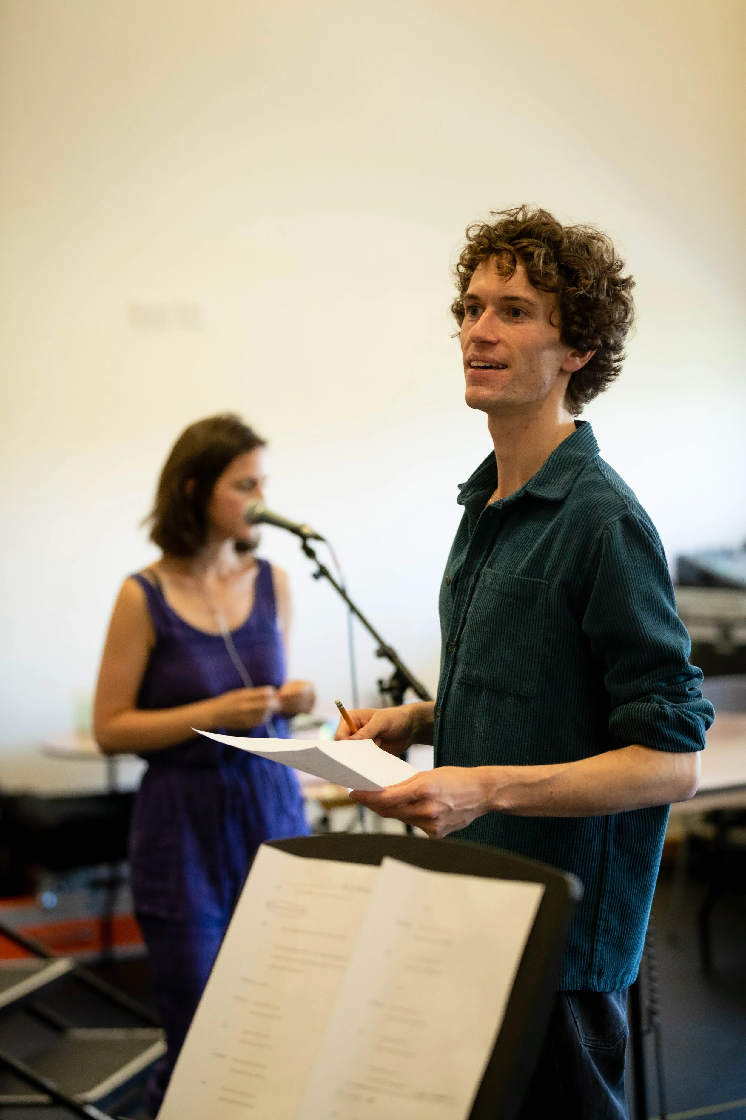 A man with curly hair holding a music score and a pencil, standing in a music rehearsal room, with a woman singing into a microphone in the background.