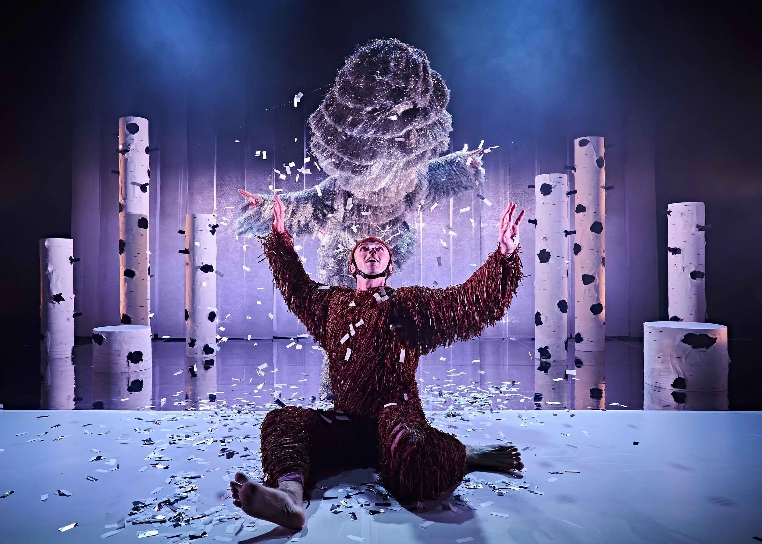 A performer in a furry costume is on their knees on a stage, with arms raised and head tilted back. They are surrounded by falling confetti and standing among tall, white, log-like pillars.