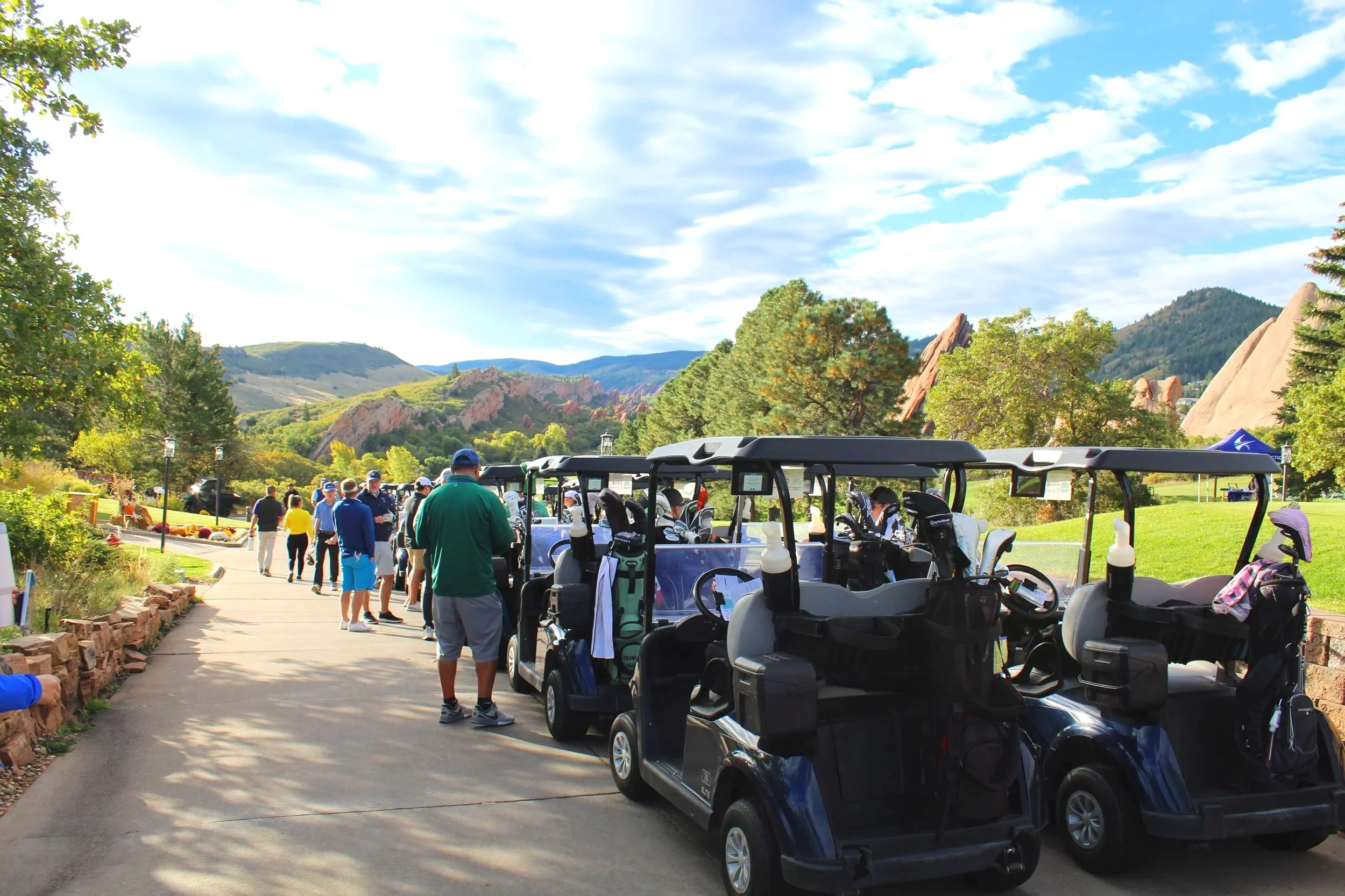 Golf carts parked along a paved pathway at a golf course with people walking and preparing to play, surrounded by green grass, trees, rocky hills, and a partly cloudy sky.