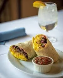 Two breakfast burritos with a side of salsa and a glass of iced beverage with lemon wedge on a white plate