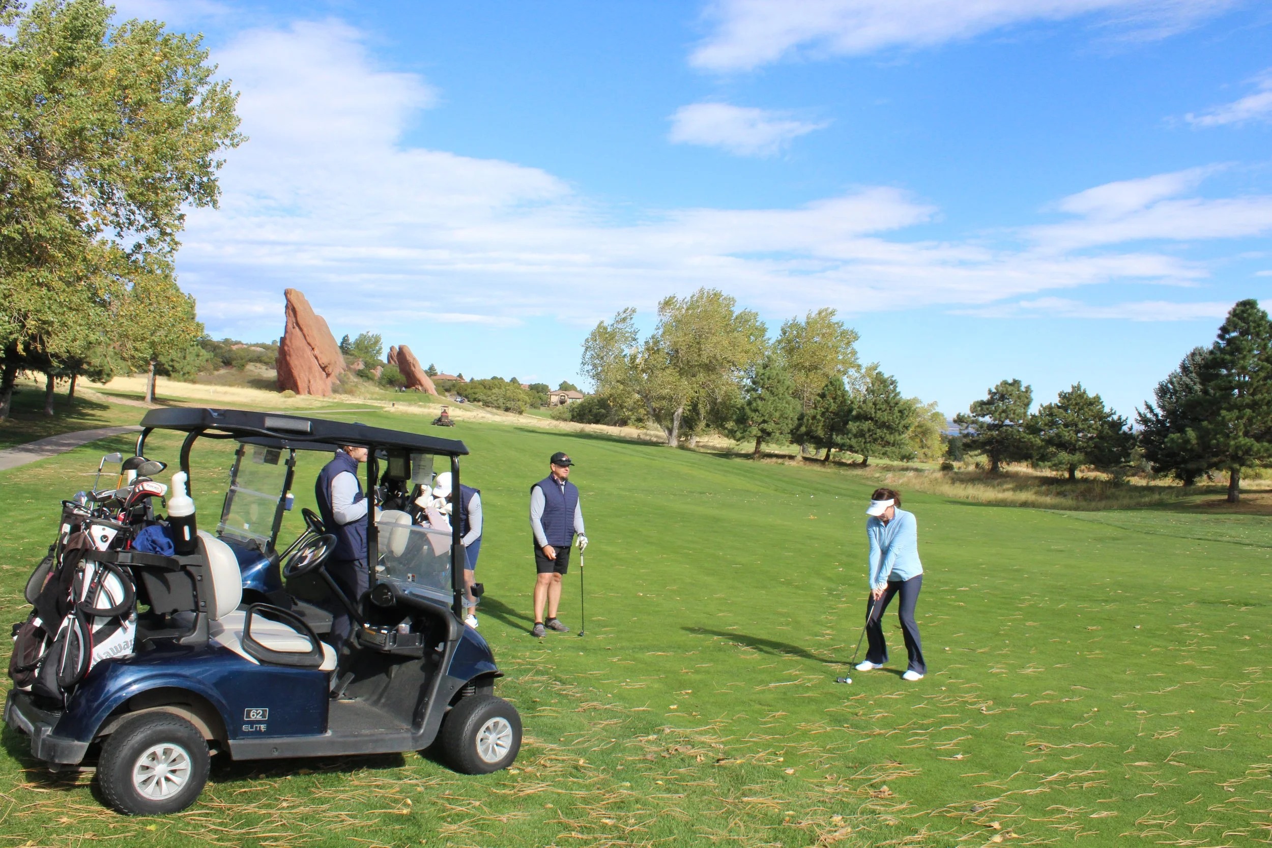A woman in a blue jacket and white visor preparing to hit a golf ball on a golf course, with four people, including a golfer, standing near her, and a golf cart with golf clubs parked on the grass under a partly cloudy sky and trees in the background