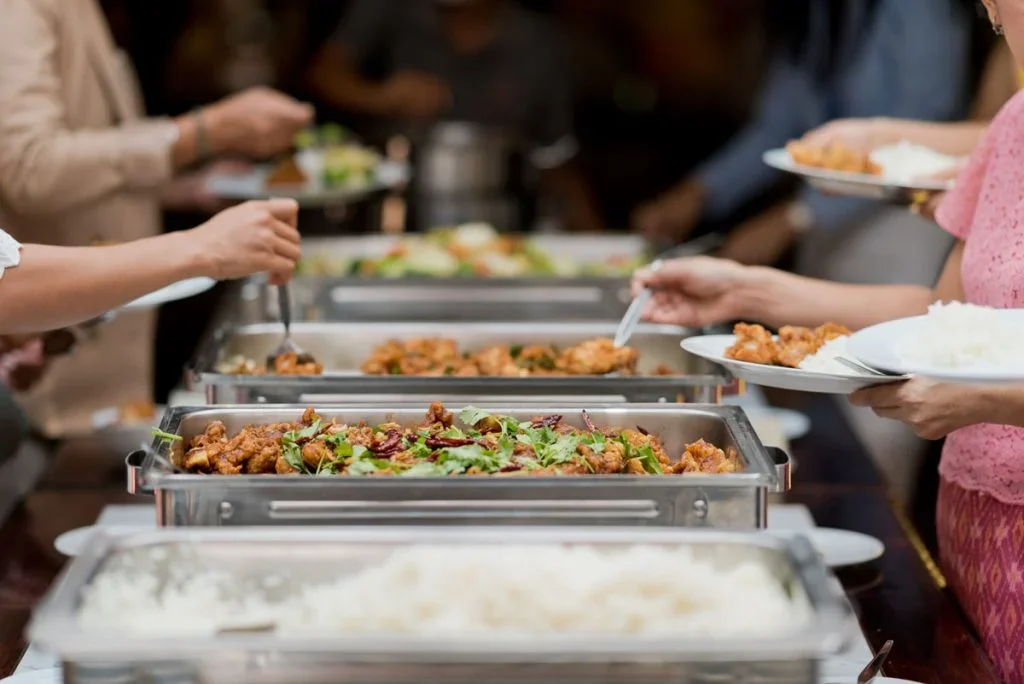 People serving themselves food from a buffet with trays of fried chicken, vegetables, and rice.
