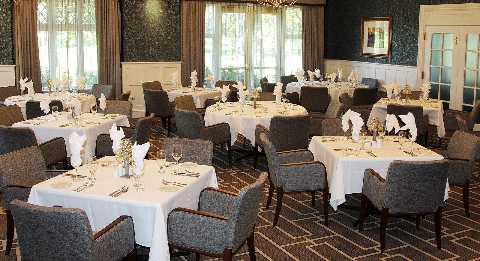 A dining room set up for a formal event, with round and rectangular tables covered in white tablecloths, each set with silverware, glassware, and white napkins folded in a decorative style. The room has large windows with curtains, dark patterned wallpaper, and framed artwork on the walls.