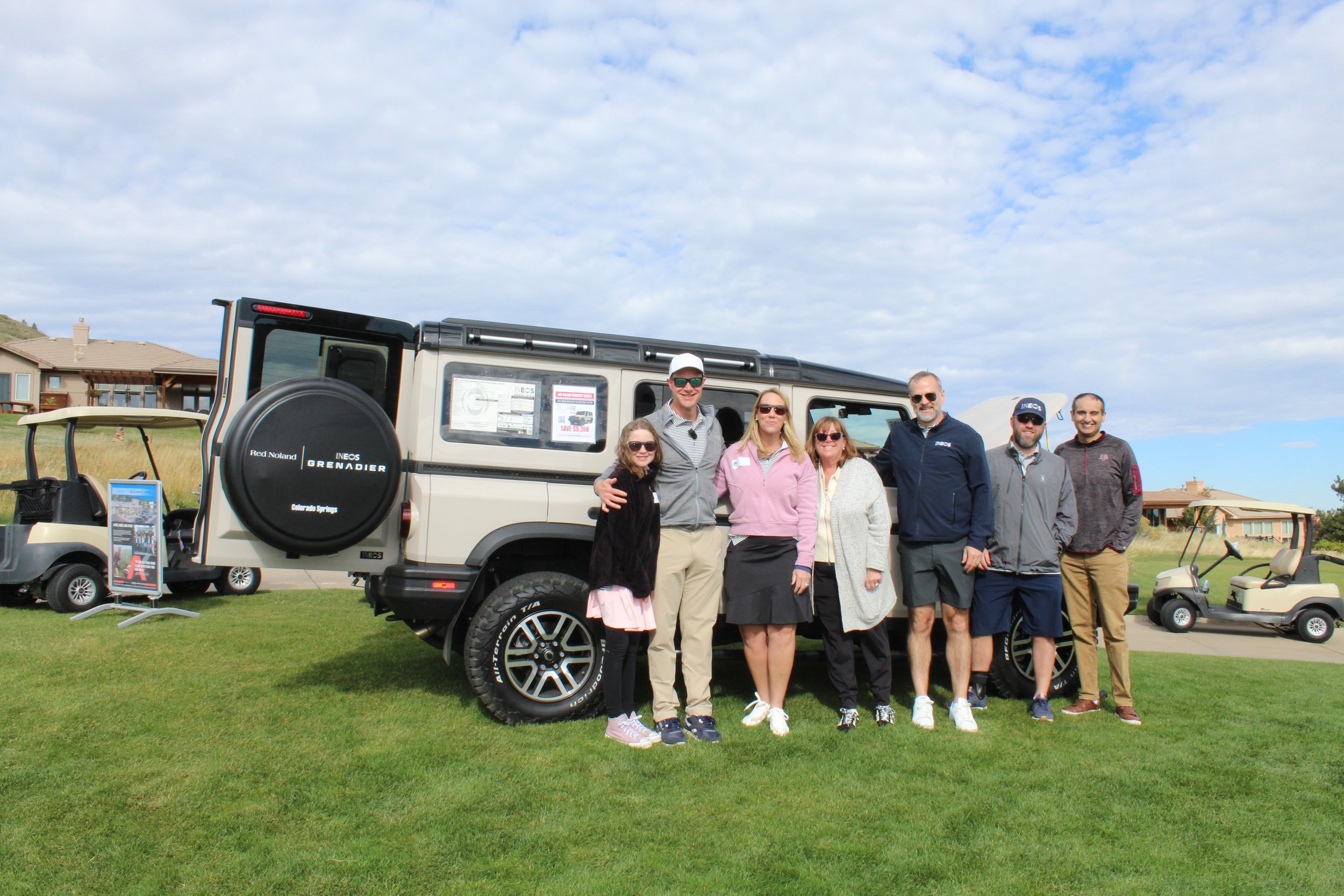 Group of seven people standing on a golf course next to a tan off-road vehicle, with golf carts and houses in the background under a partly cloudy sky.