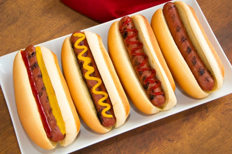 Four hot dogs in buns on a white platter, each topped with different condiments and mustard, placed on a wooden table.