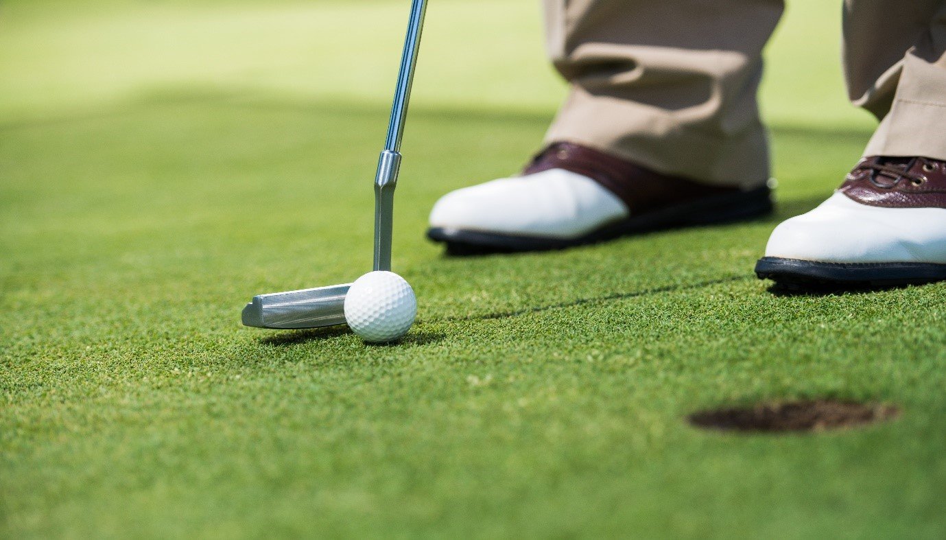 Close-up of a golfer's feet, putting with a golf club on the green, with a golf ball near the club, and a small hole in the grass