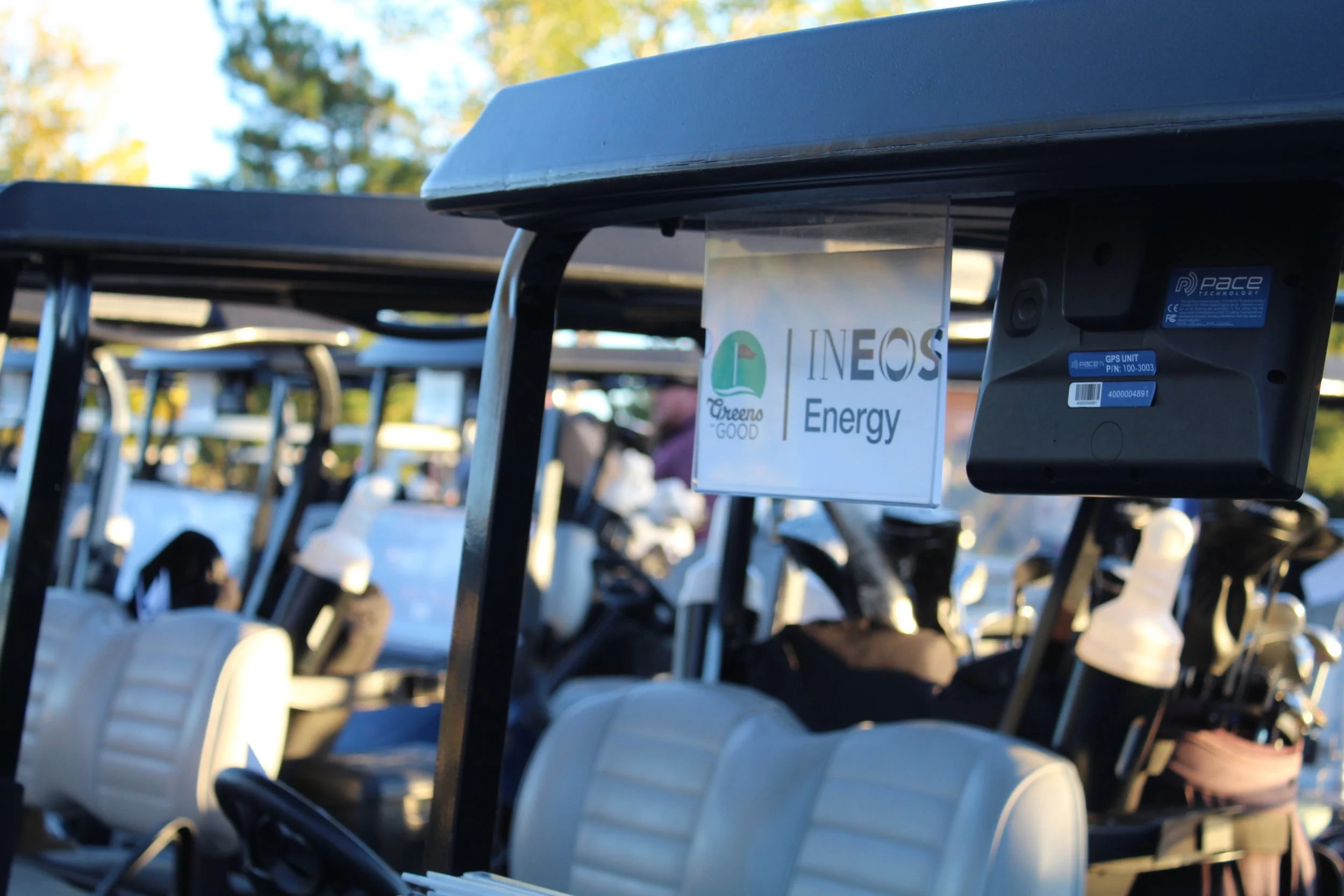 A row of golf carts parked outdoors with a sign on the front of one cart displaying the logos for Green Good, INEOS, and Energy, along with a GPS unit attached.