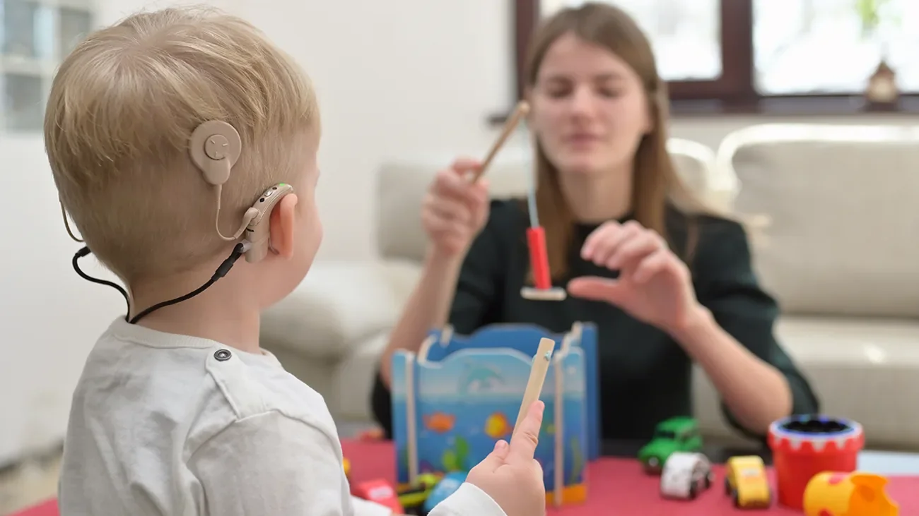 A young boy with hearing aids playing a toy fishing game with a woman, possibly his therapist, in a bright room with a window in the background.