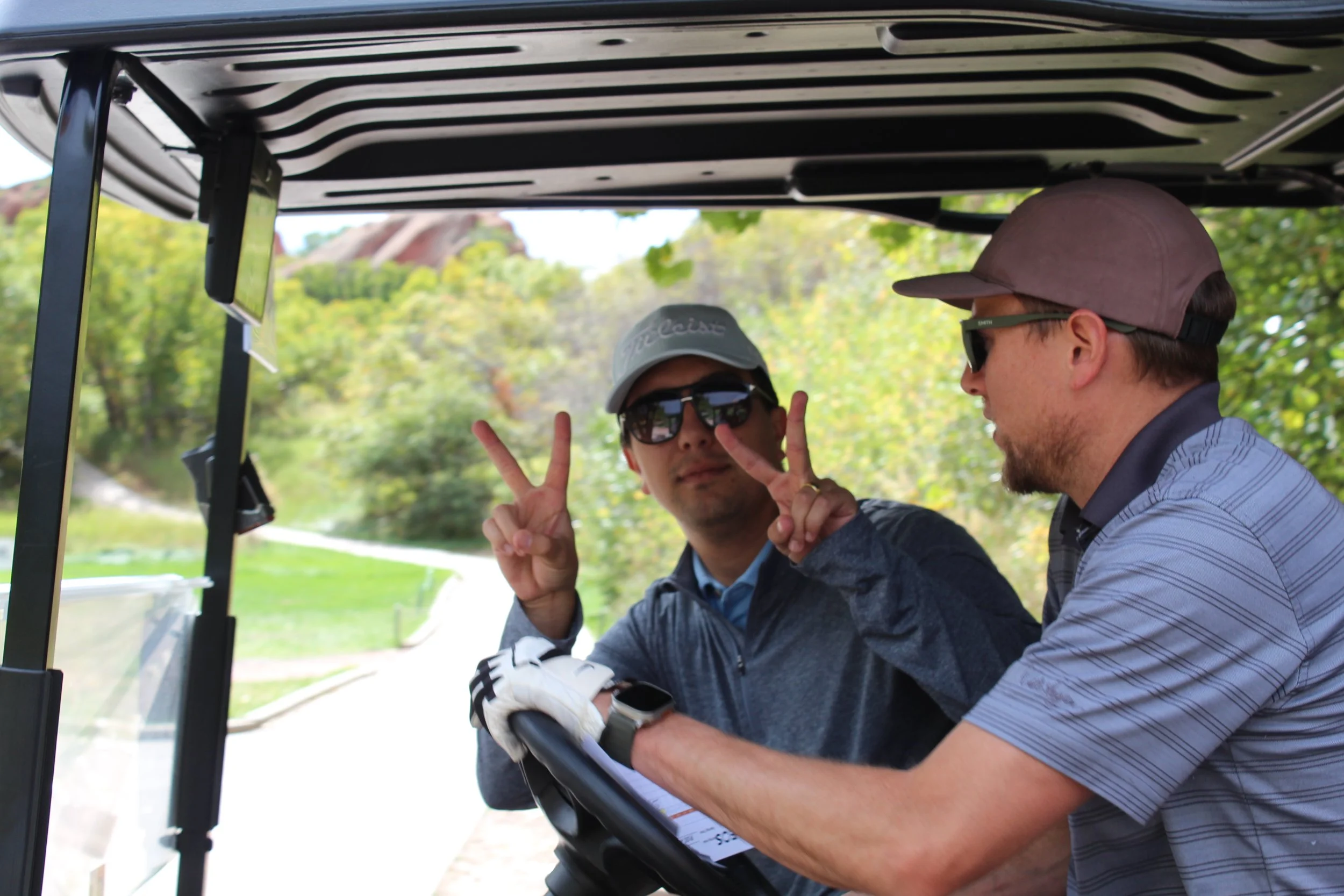 Two men sitting in a golf cart outdoors, one making peace signs with both hands and wearing sunglasses and a cap, the other wearing a cap and glasses, with trees and a golf course in the background.