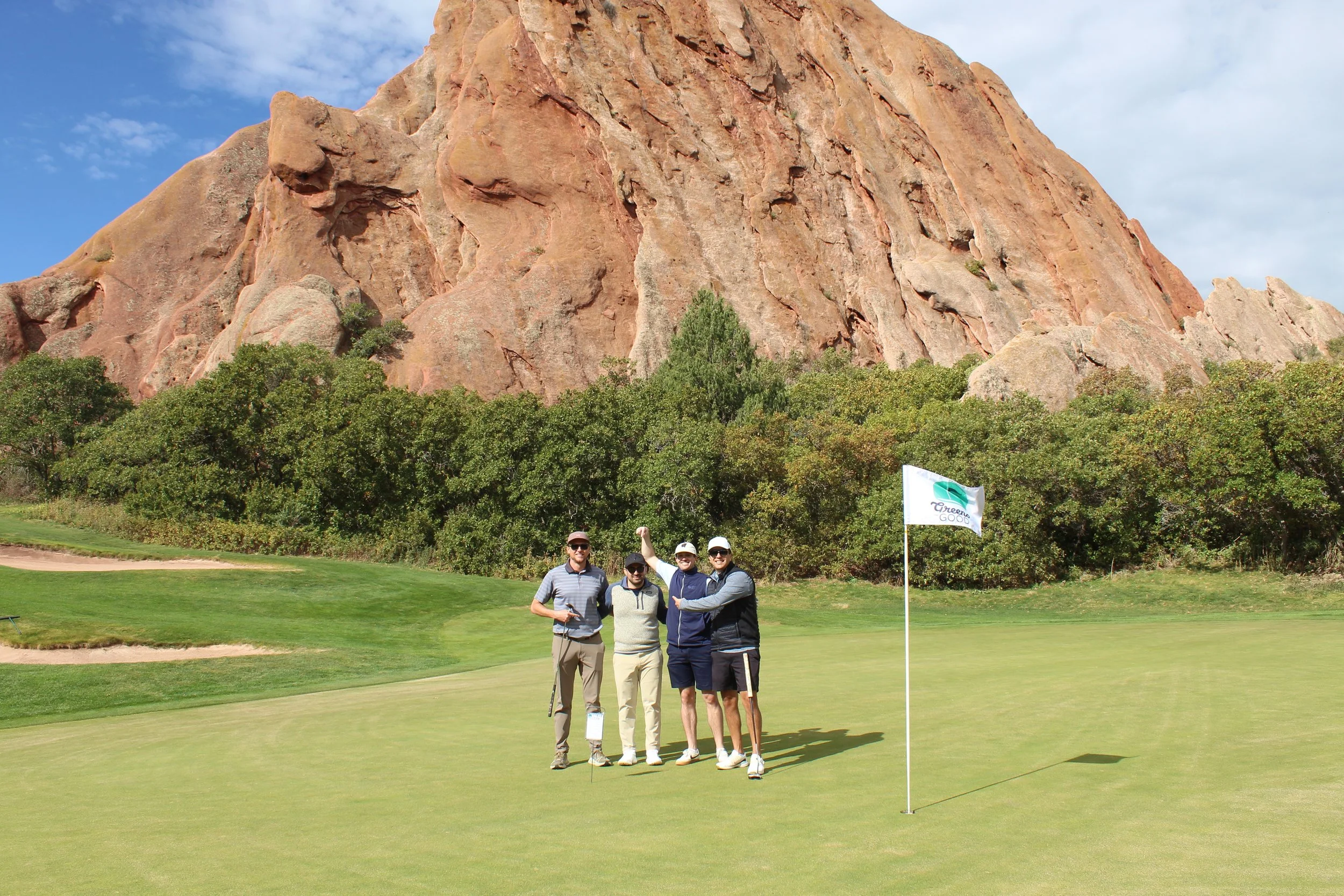 Four people standing on a golf green near a flagstick with a rocky mountain and trees in the background.