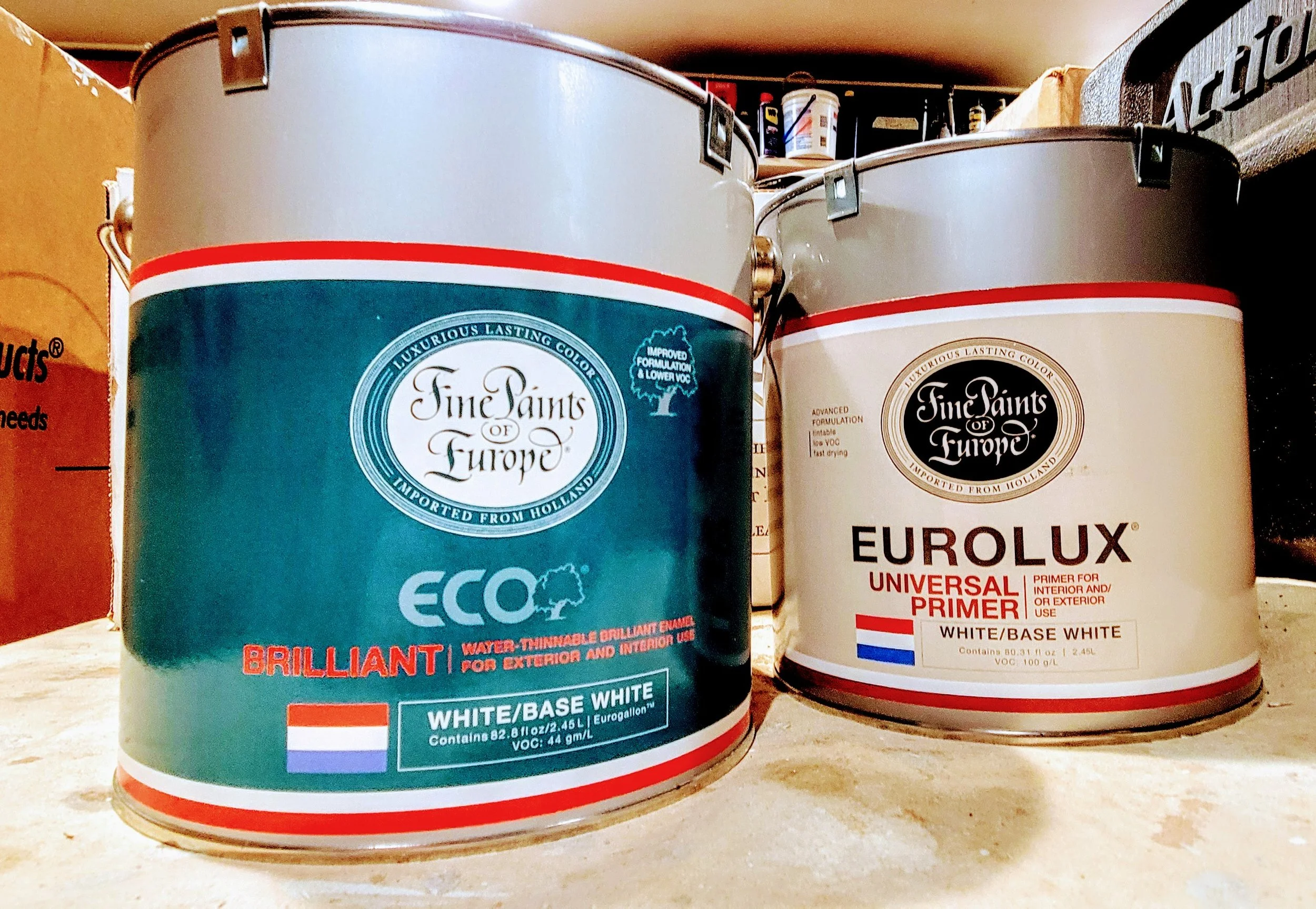 Two cans of primer paint, one labeled 'Eco Brilliant' in water-based white, the other labeled 'EuroLux Universal Primer' in white, both from Fine Paints of Europe, with Dutch flag symbols on the labels.