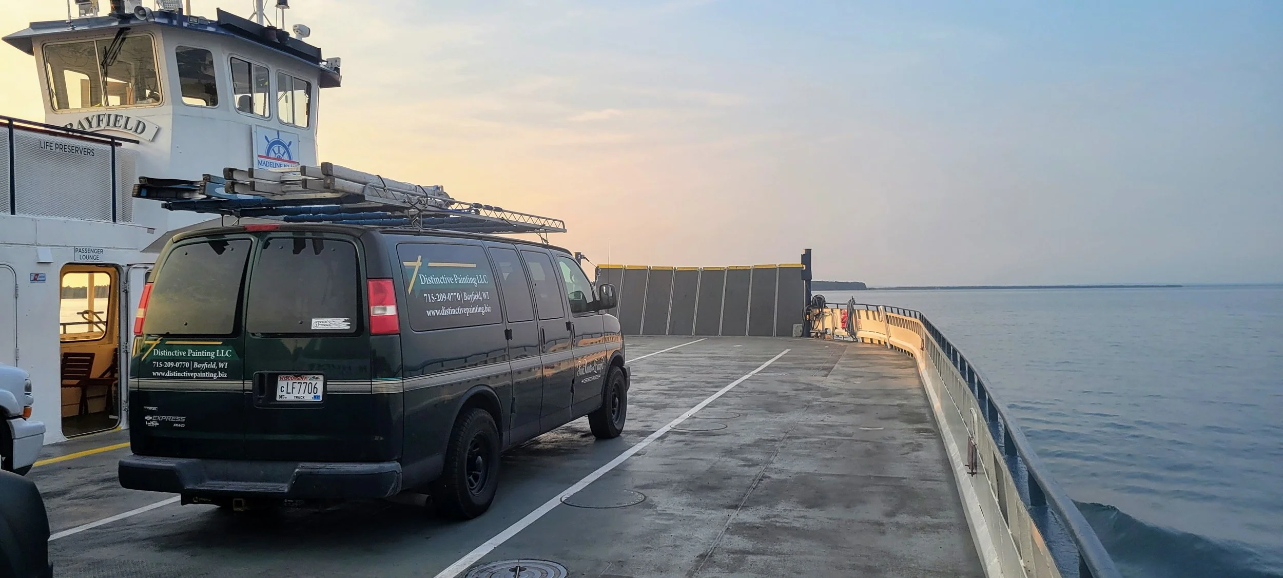 A ferry deck with a black service van labeled 'Distinctive Painting LLC' parked near the side railing. The ferry is on a body of water during sunset or sunrise, with a view of the horizon and distant shoreline. The ferry has a passenger lounge entrance and a life preserver sign visible on the upper structure.